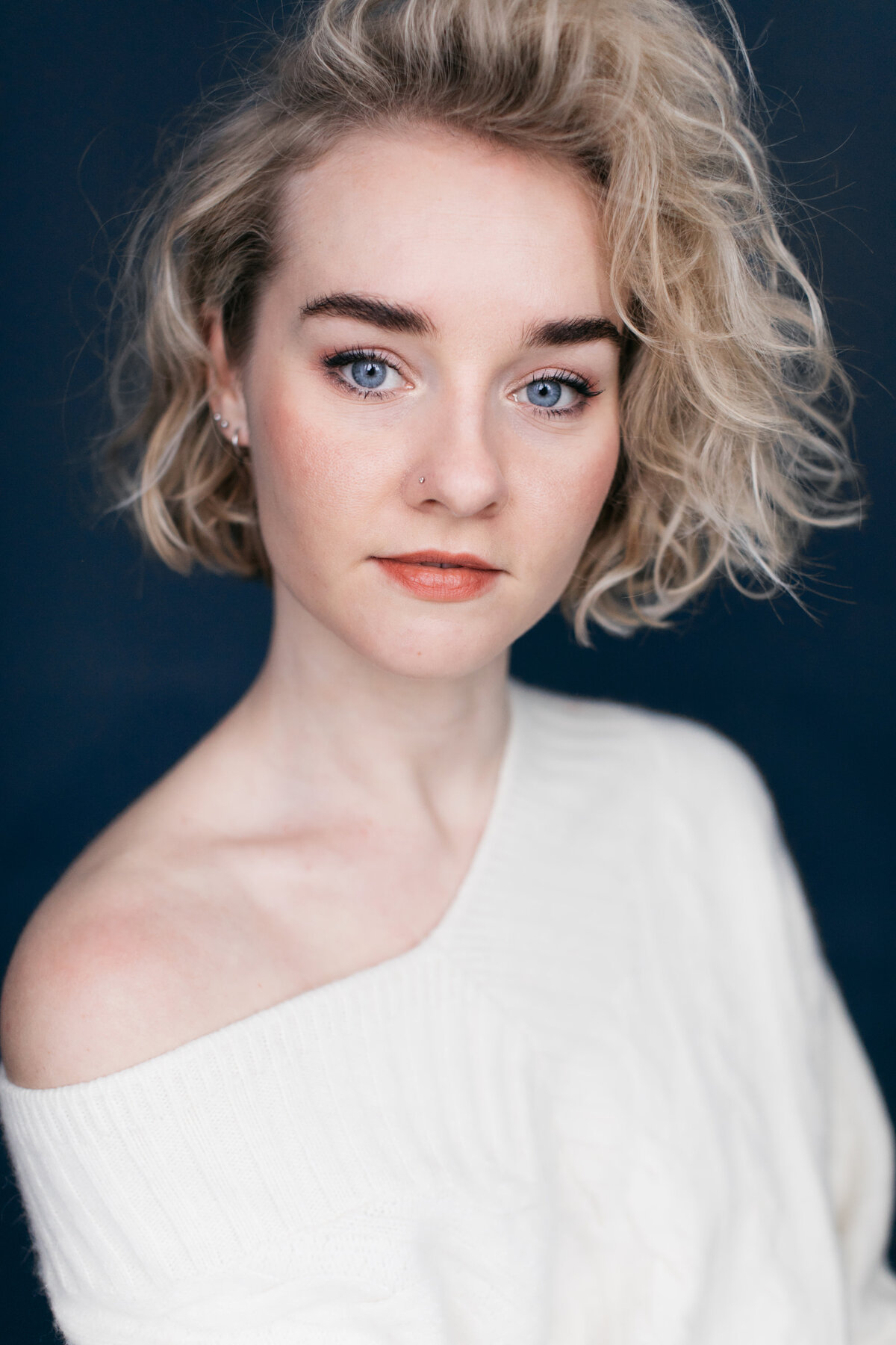 Castingfoto van een vrouw met kort blond haar in een witte top voor een blauwe achtergrond.