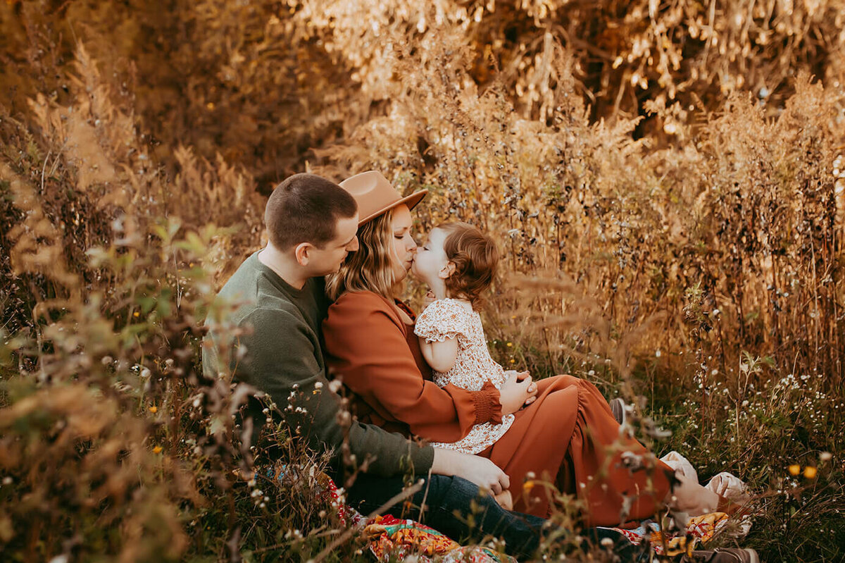 a wife sitting in her husbands lap while she holds their daughter who is kissing her mom in a field of flowers