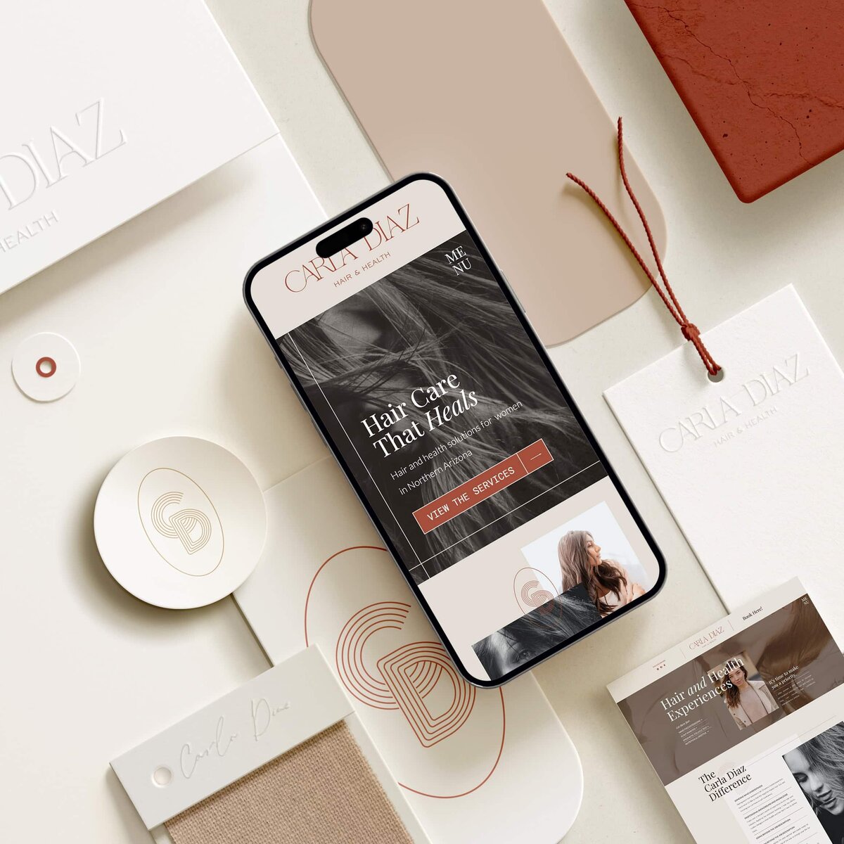 A smartphone displaying a hair care website rests on a beige surface, surrounded by various branded stationery items including business cards, envelopes, and textured paper—elements that beautifully showcase cohesive branding and website design services.