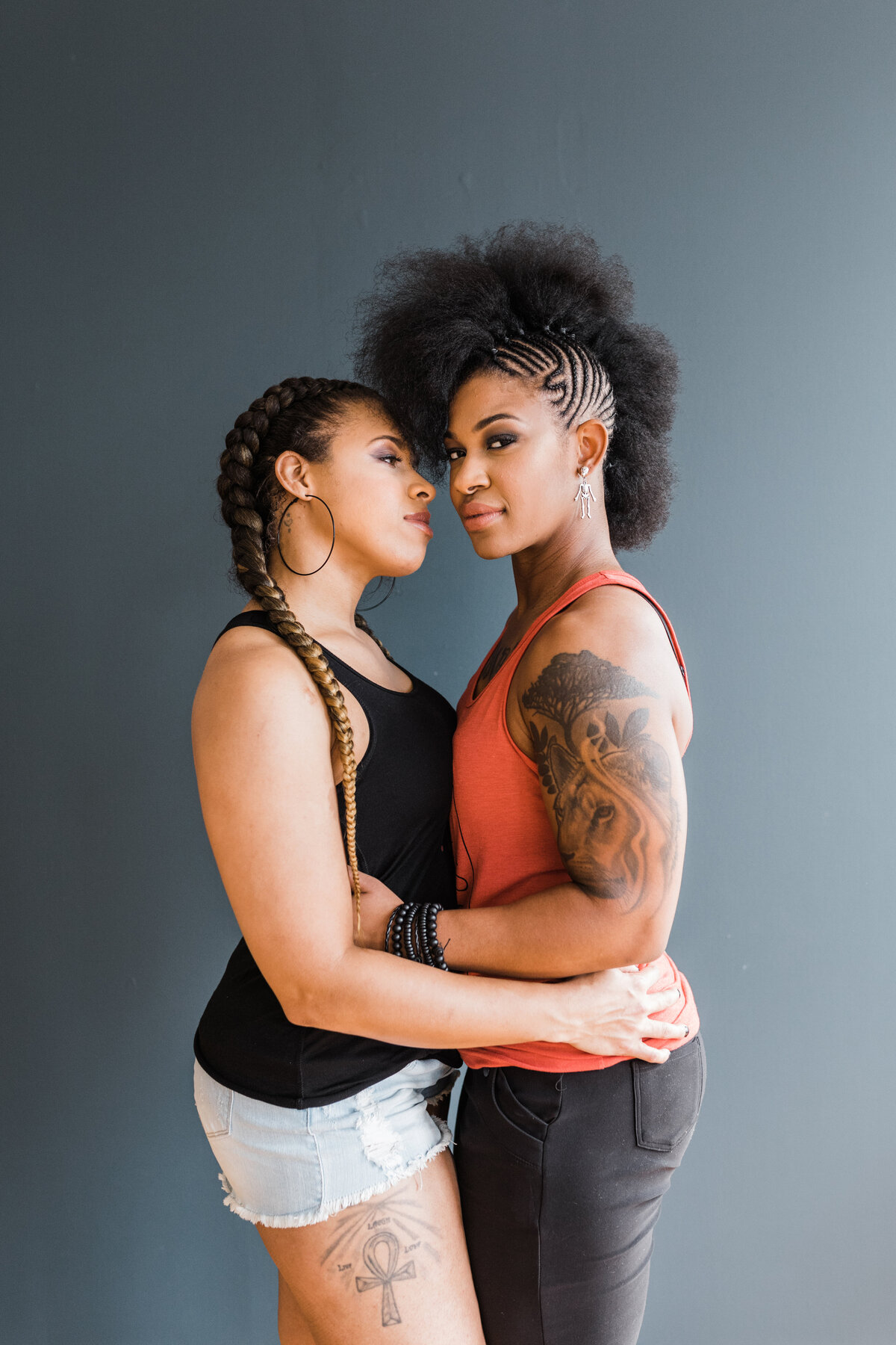 Two women posing intimately during their studio engagement photoshoot in Dallas, Texas. They are both holding each other close in front of a dark background. The woman on the left is wearing a black tank top with cutoff denim shorts. The woman on the right is wearing an orange tank top with black jeans. Both have intricate braids and tattoos.