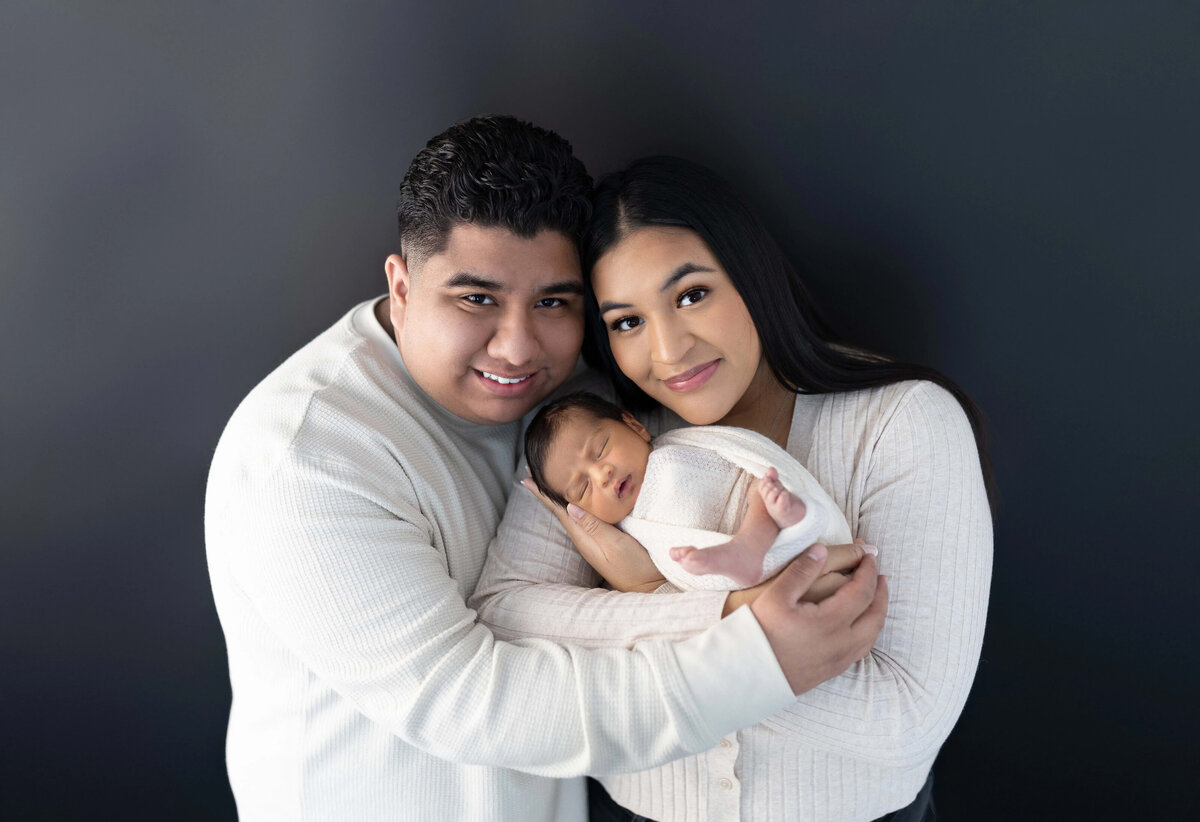 Parents snuggling newborn wrapped in white against dark backdrop - studio newborn photography Minneapolis