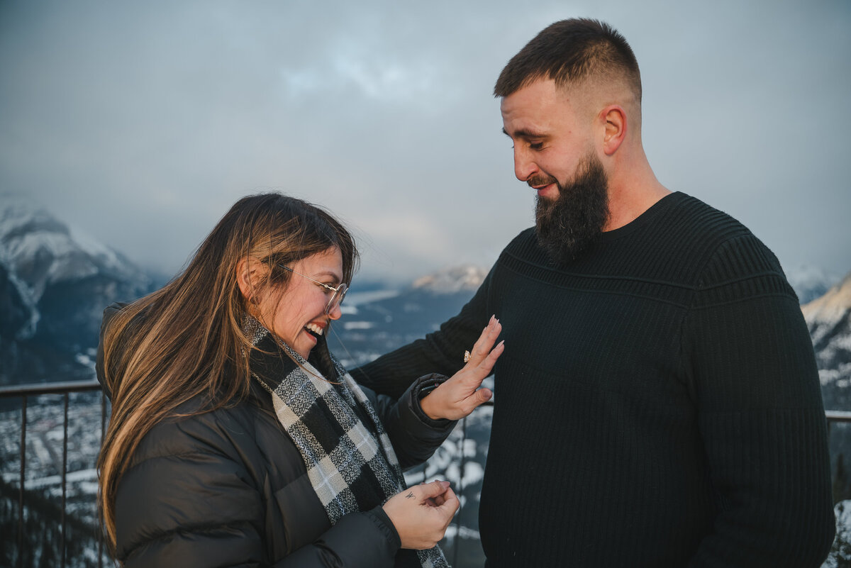 Woman reacts to surprise engagement ring while standing with fiancé at a scenic viewpoint in Banff.