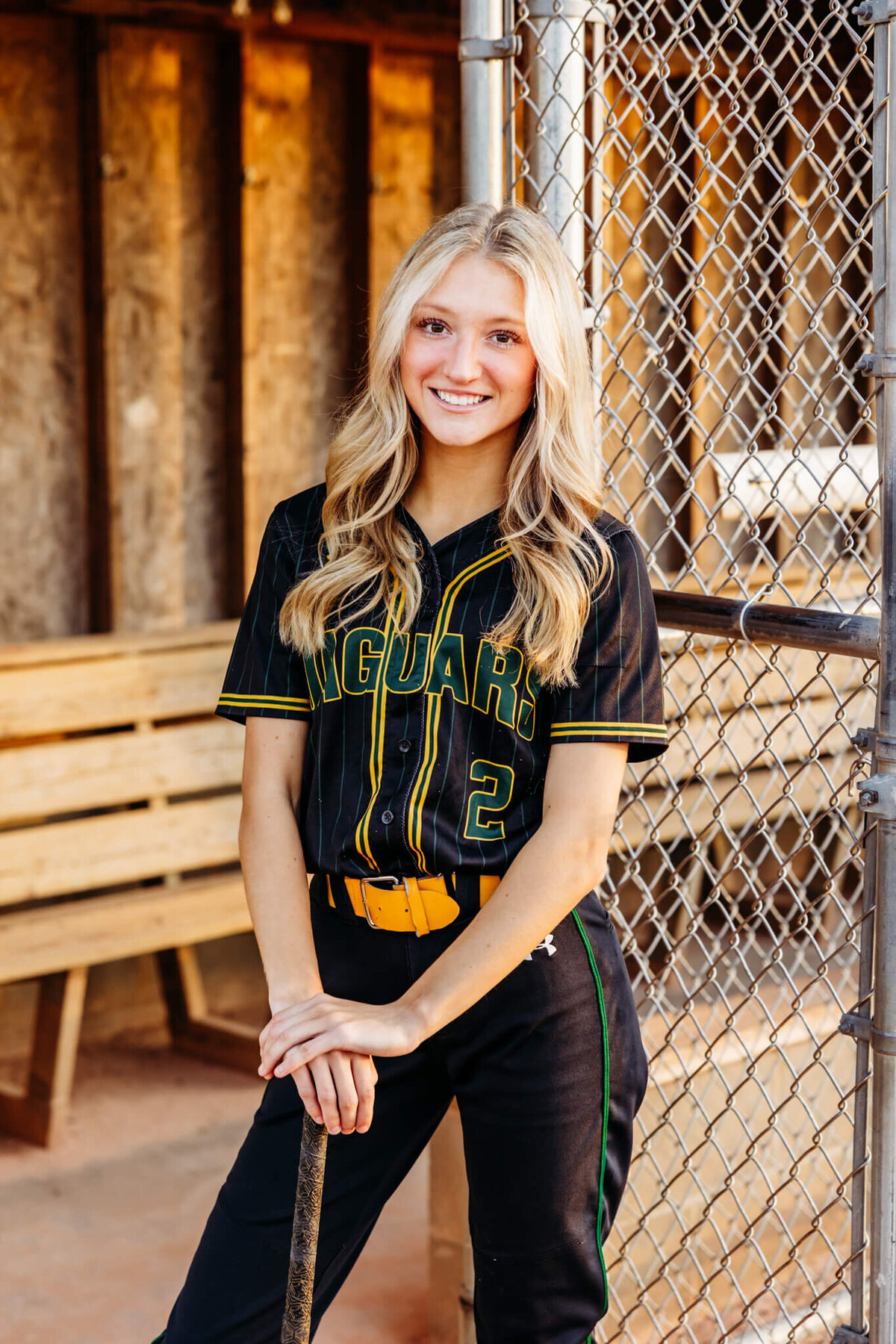 pretty teenage girl in her softball uniform resting her hands on her bat as she smiles for her senior photography session.