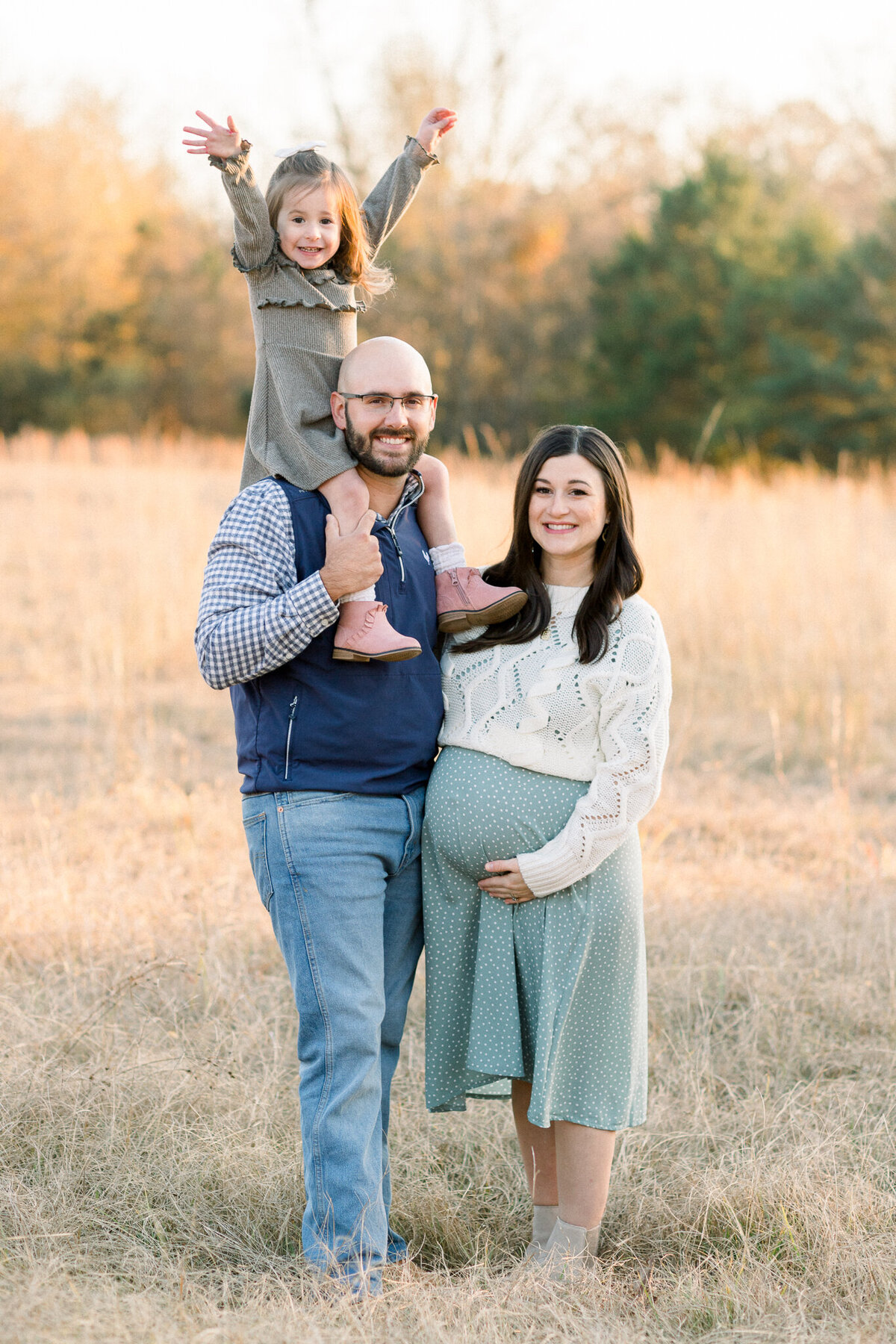 Mom to be wearing long light blue dress with white sweater and posing with husband and little girl in olive green dress in open field by central Mississippi maternity photographer.