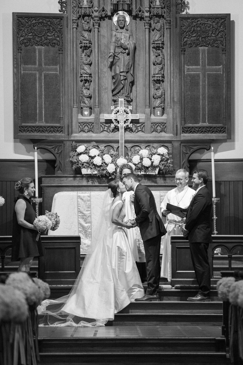Black and white photo of a wedding ceremony in a church. This is where the photo was taken at The Hamptons, New York, waterfront estate tented wedding by photographer Bonnie Sen. The bride and groom kiss at the altar, flanked by two attendants. A priest stands behind them, under a statue and crucifix surrounded by flowers. Videographers: East West Films.