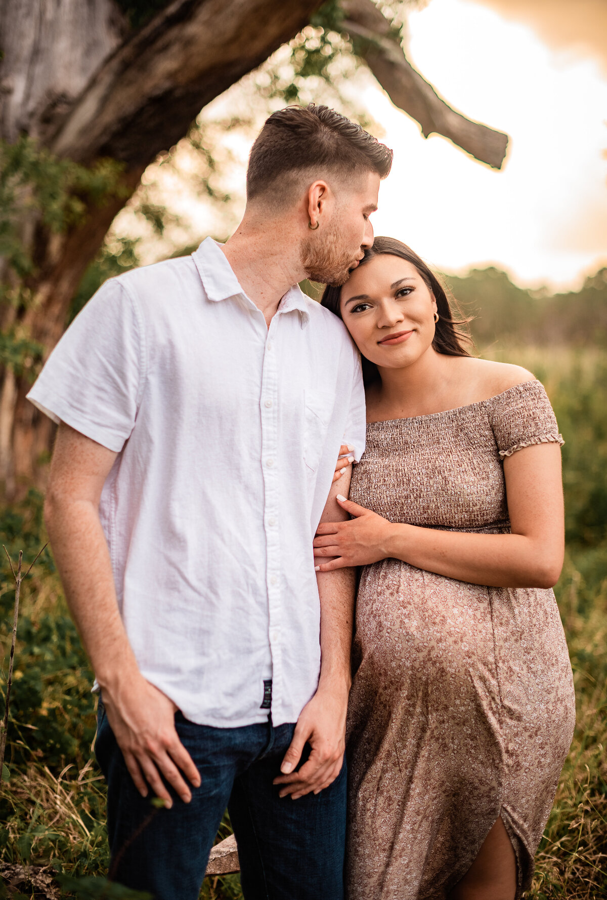 An expectant mother hugs her boyfriend's arm while he kisses the top of her head.
