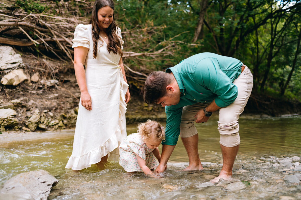 Family photo In a river, the woman is smiling in a long white dress and is watching the girl and man play with water. The girl sits inside the water in a white dress and the man is crouched touching the water in a green shirt and beige trousers. Behind them are some roccas and some trees with branches