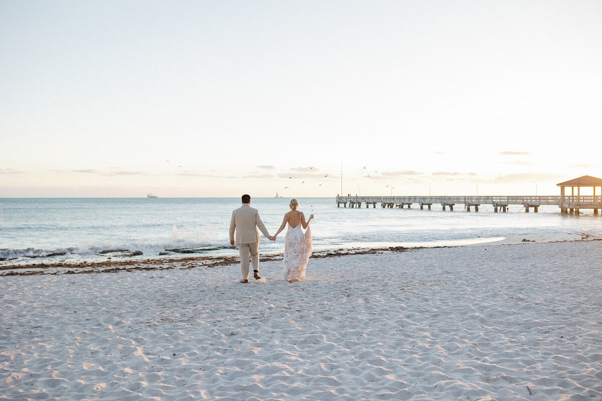 married couple standing on beach with back to camera looking at the water with a pier extending from the right