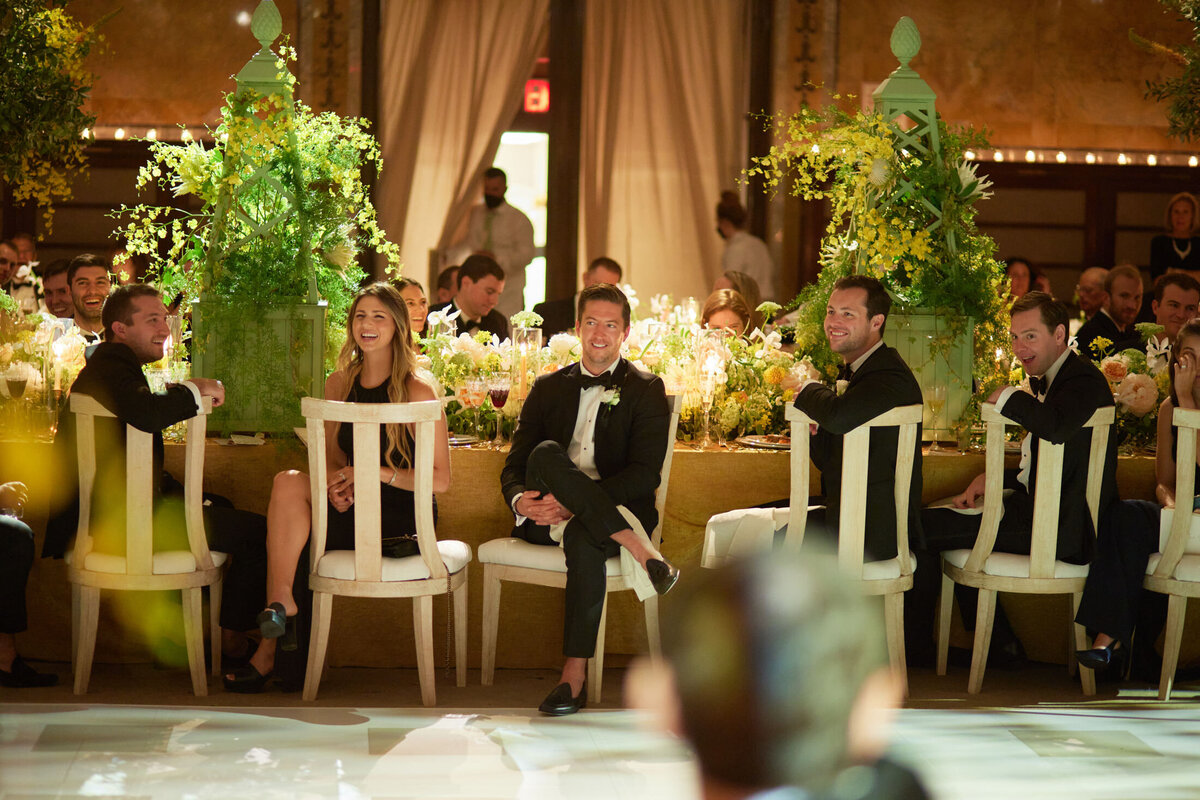 The photo was taken at The New York Public Library in New York City by photographer Shawn Connell, capturing a group of people in formal attire seated at a long, elegantly decorated dining table. The setting suggests an event or celebration with floral arrangements and ambient lighting. The focus is on five individuals at the front, smiling and engaged in conversation. Aaron Navak Films can be added as videographers for this event.
