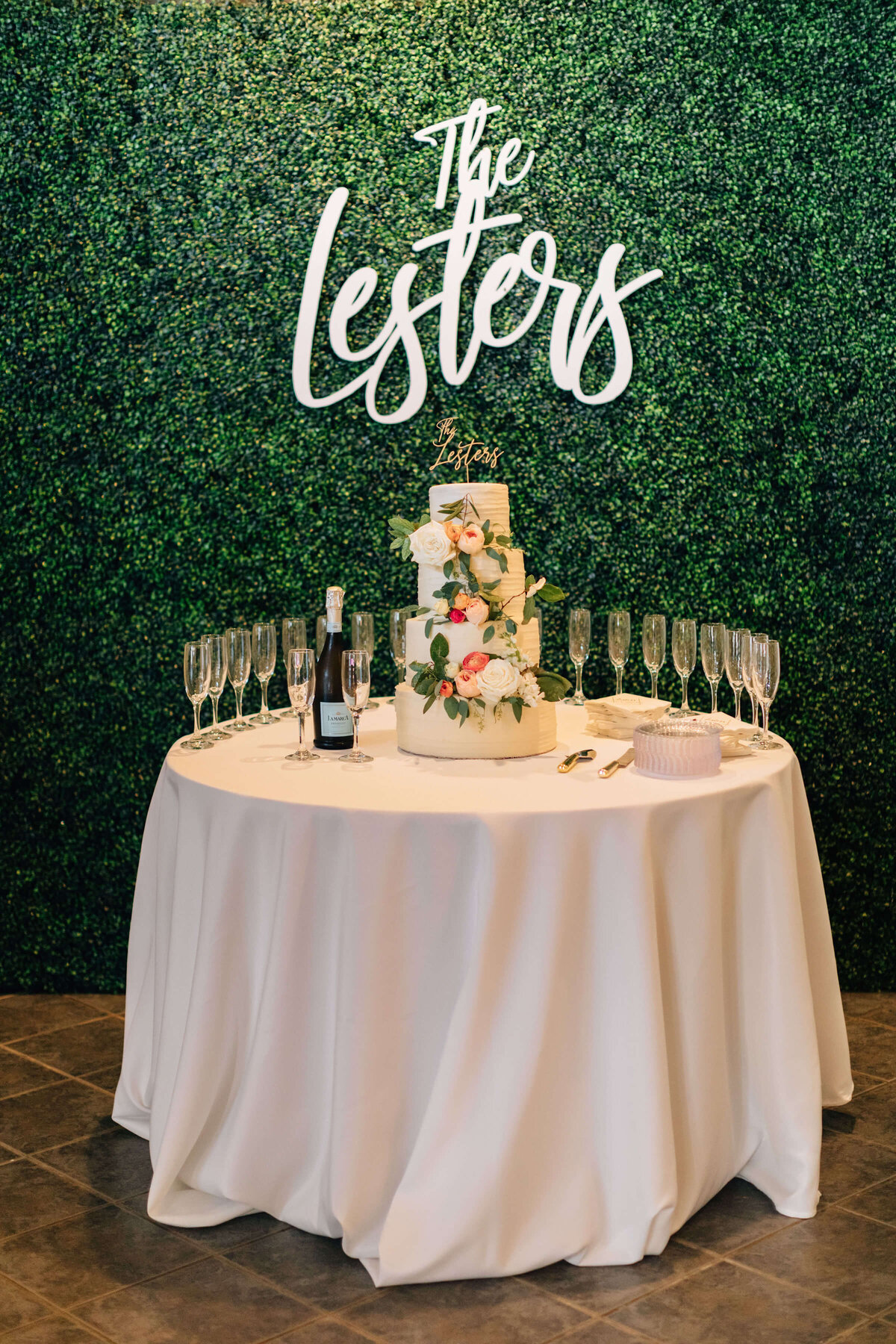 A round table with a white cloth holds a two-tiered floral cake and glasses. Behind it is a green leafy wall with The Lesters sign in white script at the top.