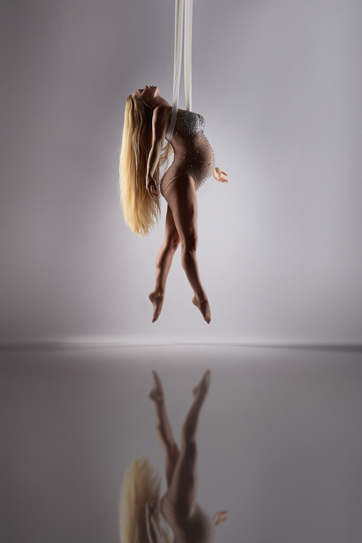 Woman gracefully suspended from white aerial silks, wearing a sparkling sheer bodysuit. Her long blonde hair cascades down as she elegantly poses in midair, reflecting on the polished surface below. Maternity photography by Lindsay Walden Photography.
