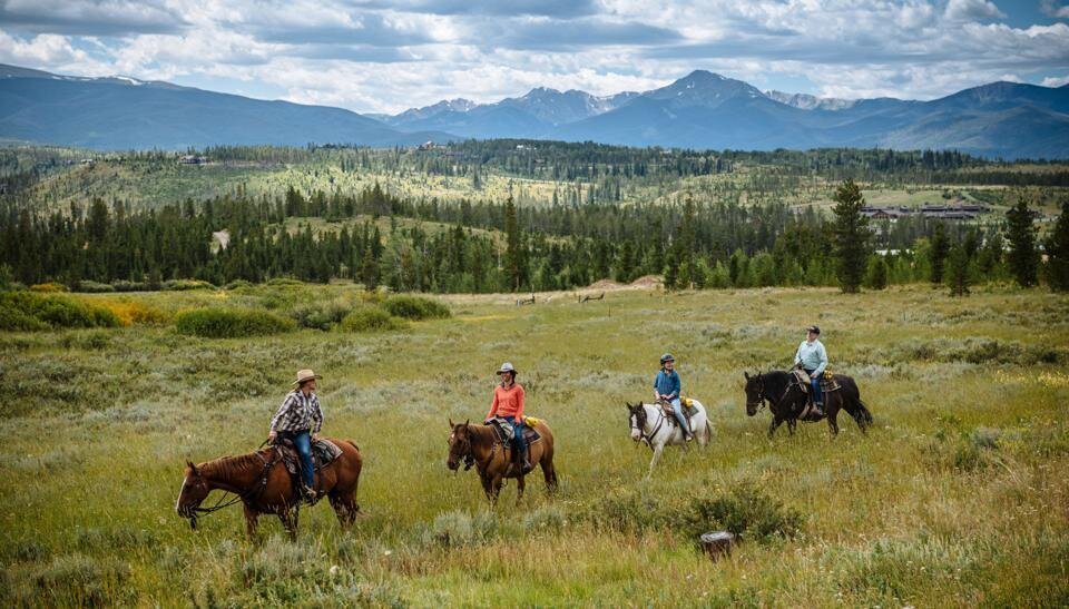 People riding on horse back through grassy fields with the mountains in the background