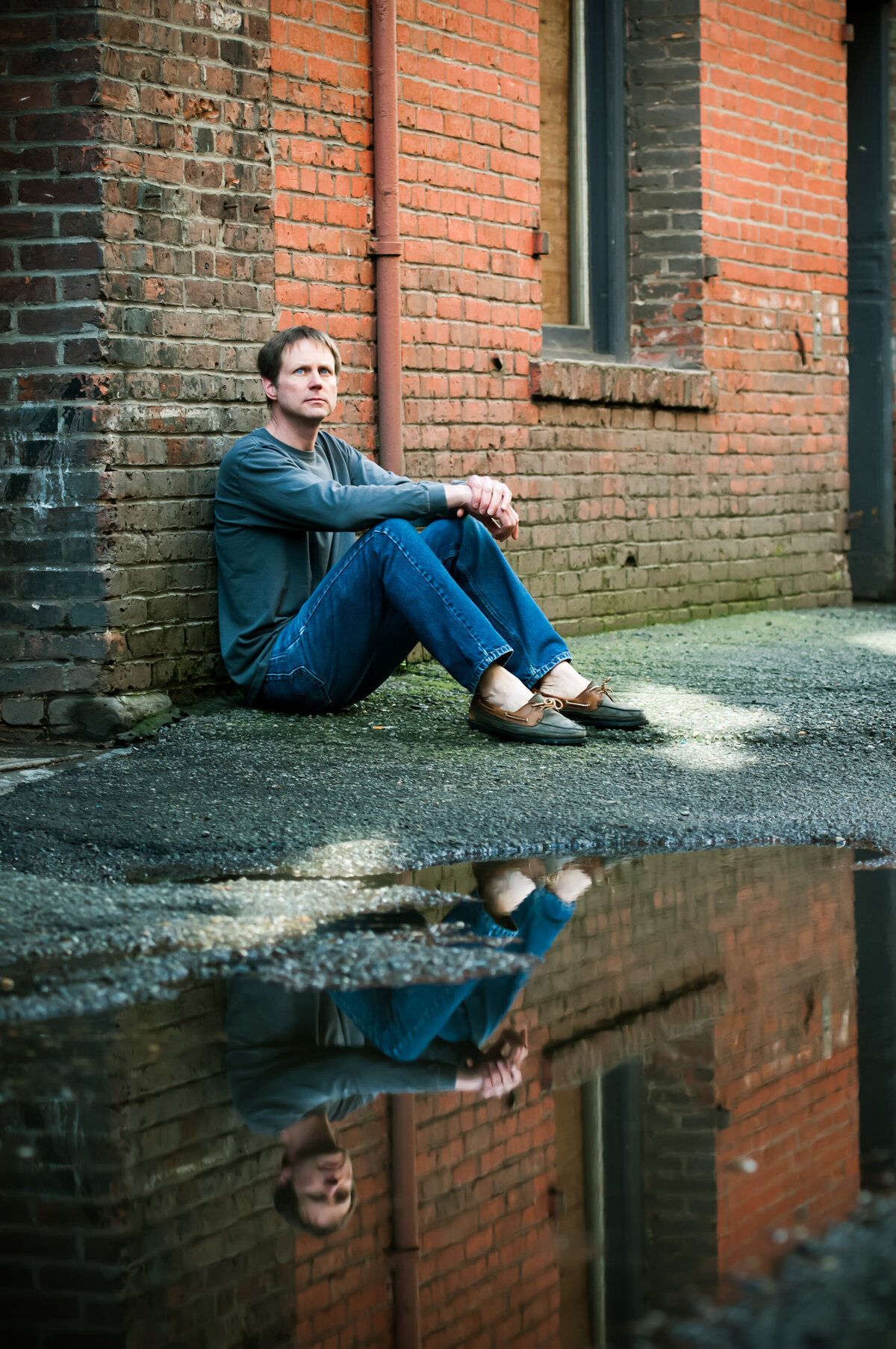 A person sitting on the ground in an alley with their reflection showing in a puddle in front of them