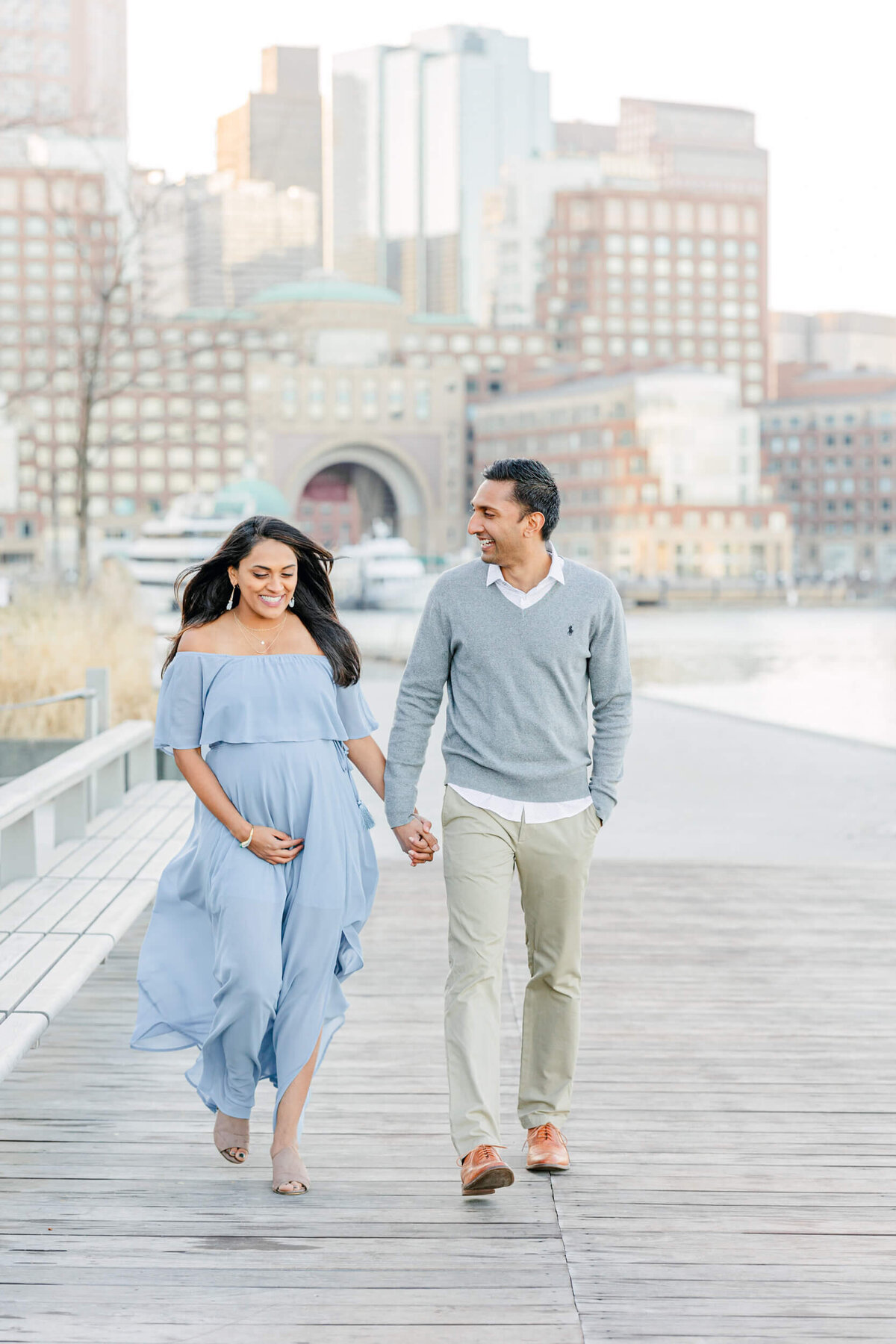 Pregnant woman and her husband hold hands and smile together as they walk in the Boston Seaport district