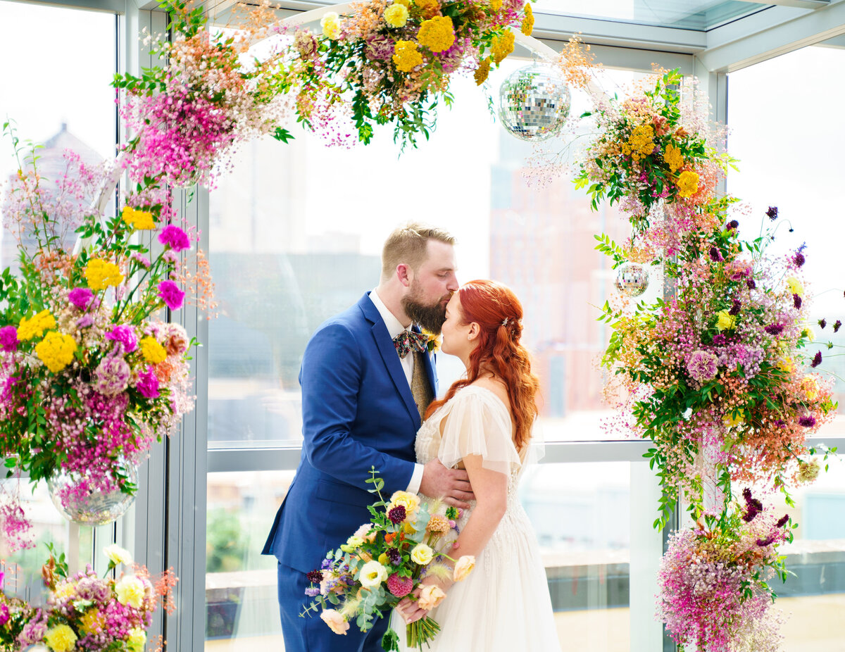 Groom kisses his bride on the forehead under a colorful, whimsical floral arch with disco balls with the skyline behind them at their wedding at North 4th Corridor (The Revery) in Columbus, Ohio.