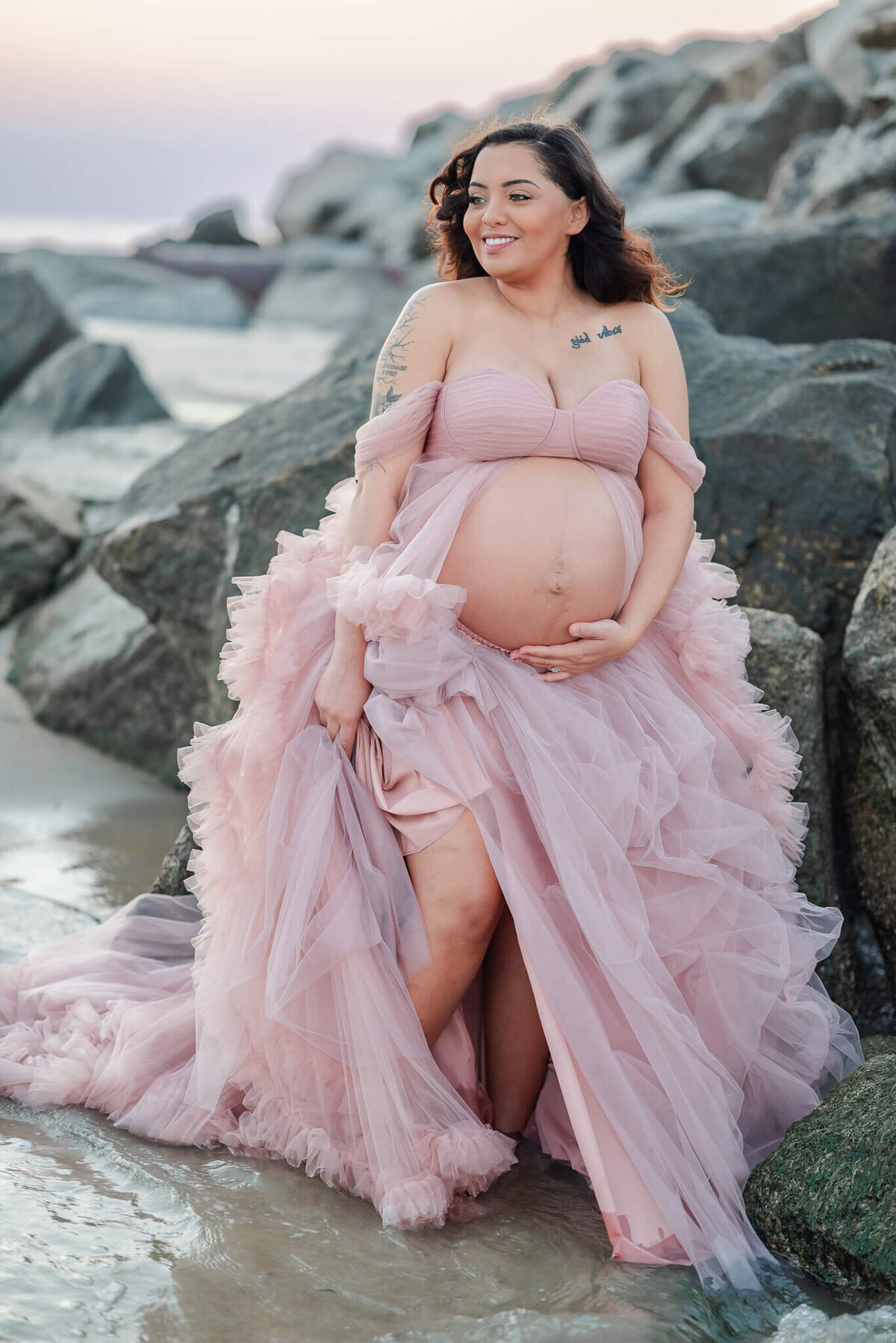 A pregnant woman stands near some rocks on the beach. She holds her pink tulle skirt up while a wave comes in.