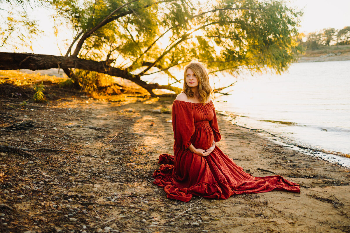 Pregnant woman sitting on the sand in front of a lake and a tree. She is wearing a long red dress, and is looking to the left side while holding her belly