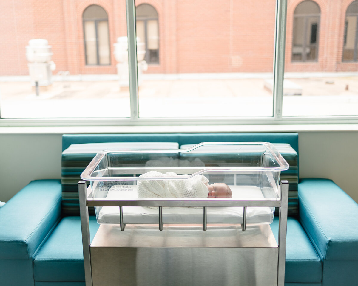 Photo of newborn baby in the hospital, he is lying with a white blanket. Behind is a light blue sofa and a large window.