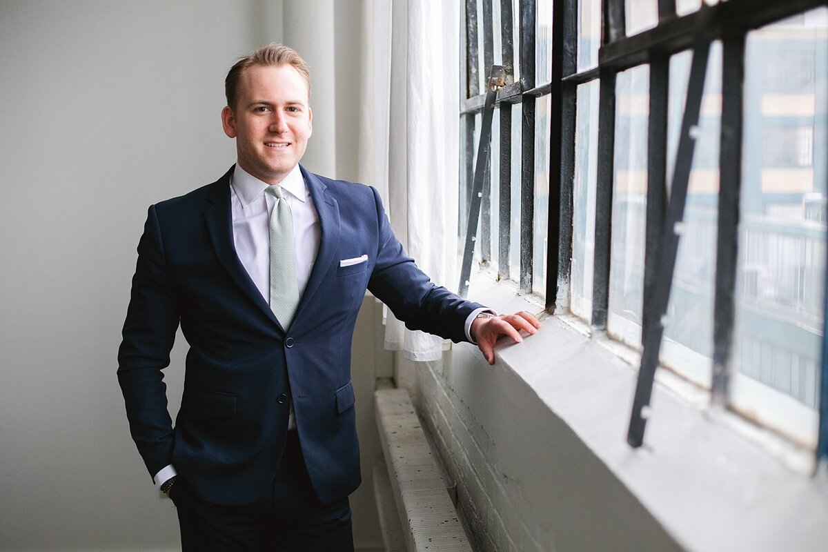 a-man-in-a-blue-blazer-and-white-button-down-smiling-by-a-large-industrial-window-with-soft-natural-light-in-indianapolis