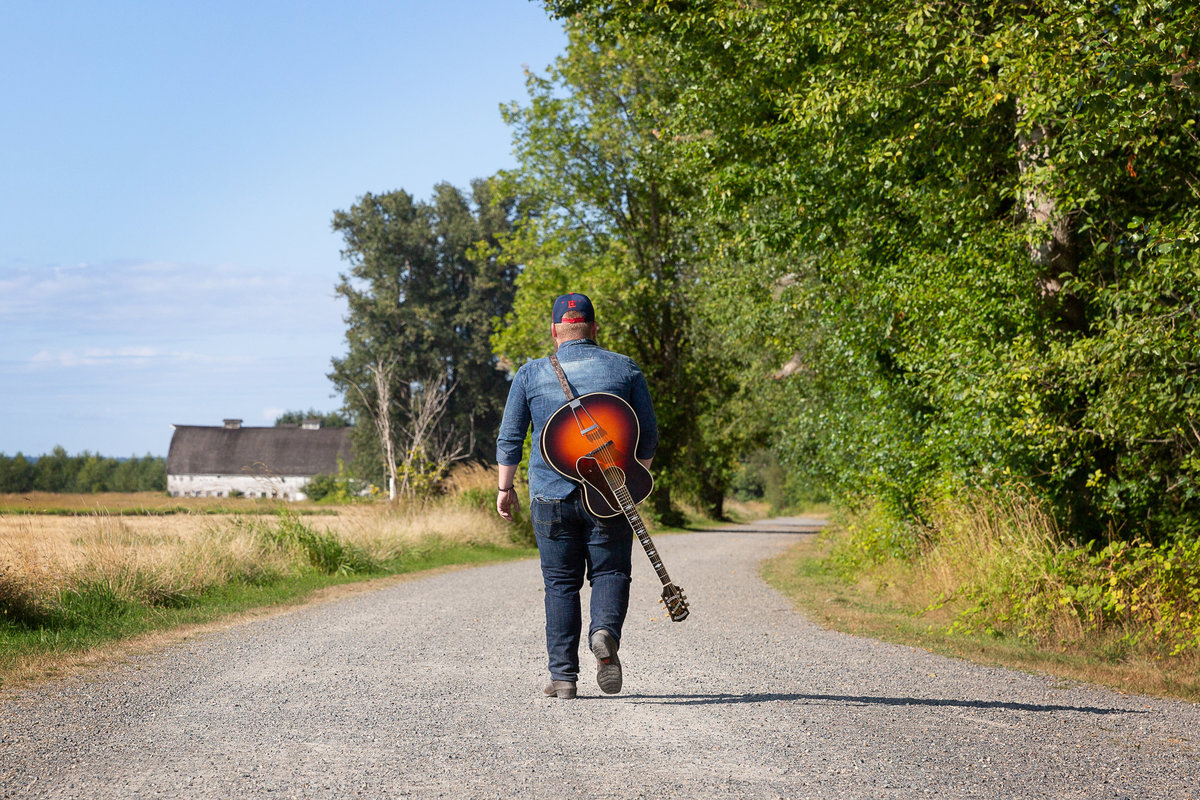 A senior guy walks away on a dirt road with a guitar slung over his back