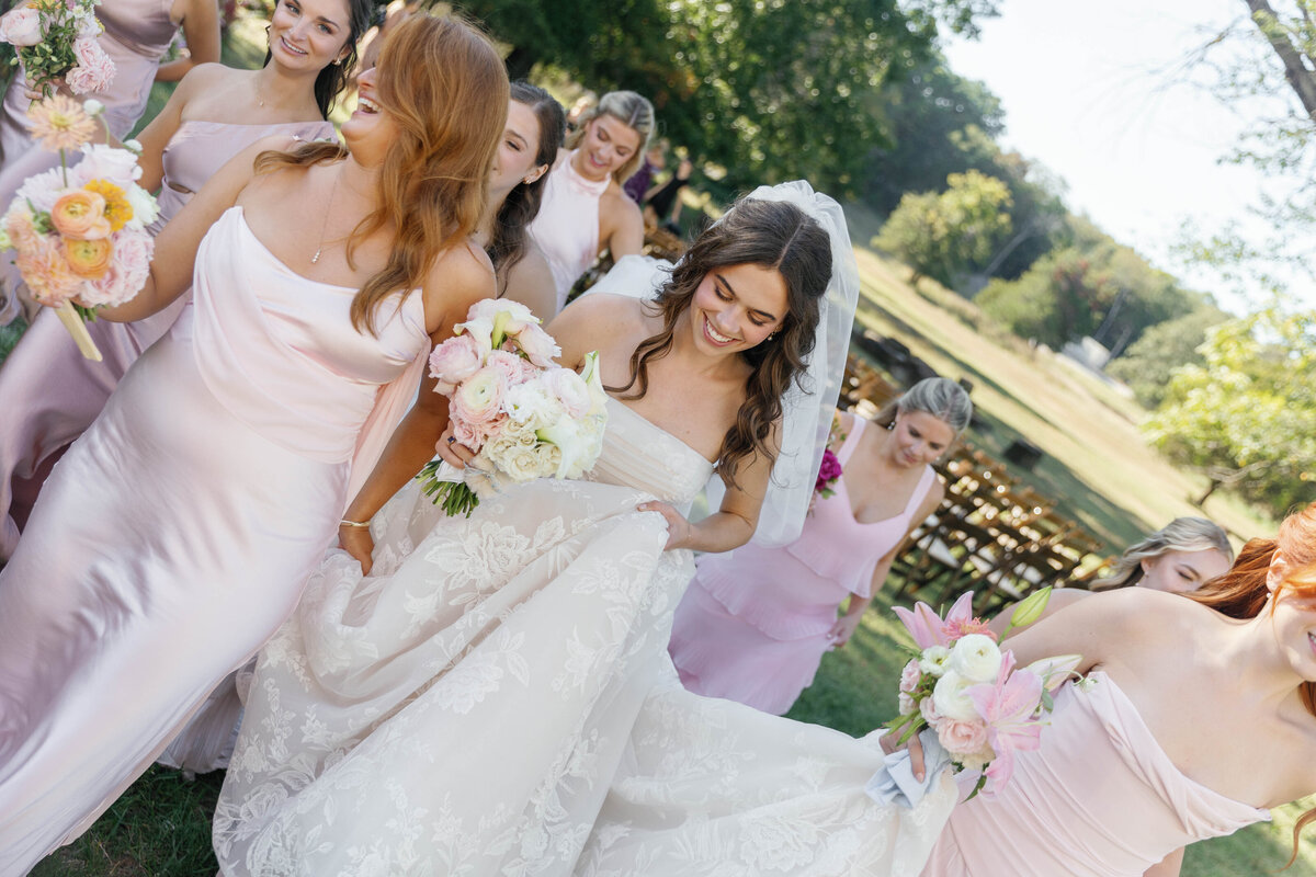 Bride walking with bridesmaids in soft blush dresses holding pastel floral bouquets featuring roses, dahlias, and lilies during an outdoor Arkansas wedding, showcasing romantic garden-style floral design.
