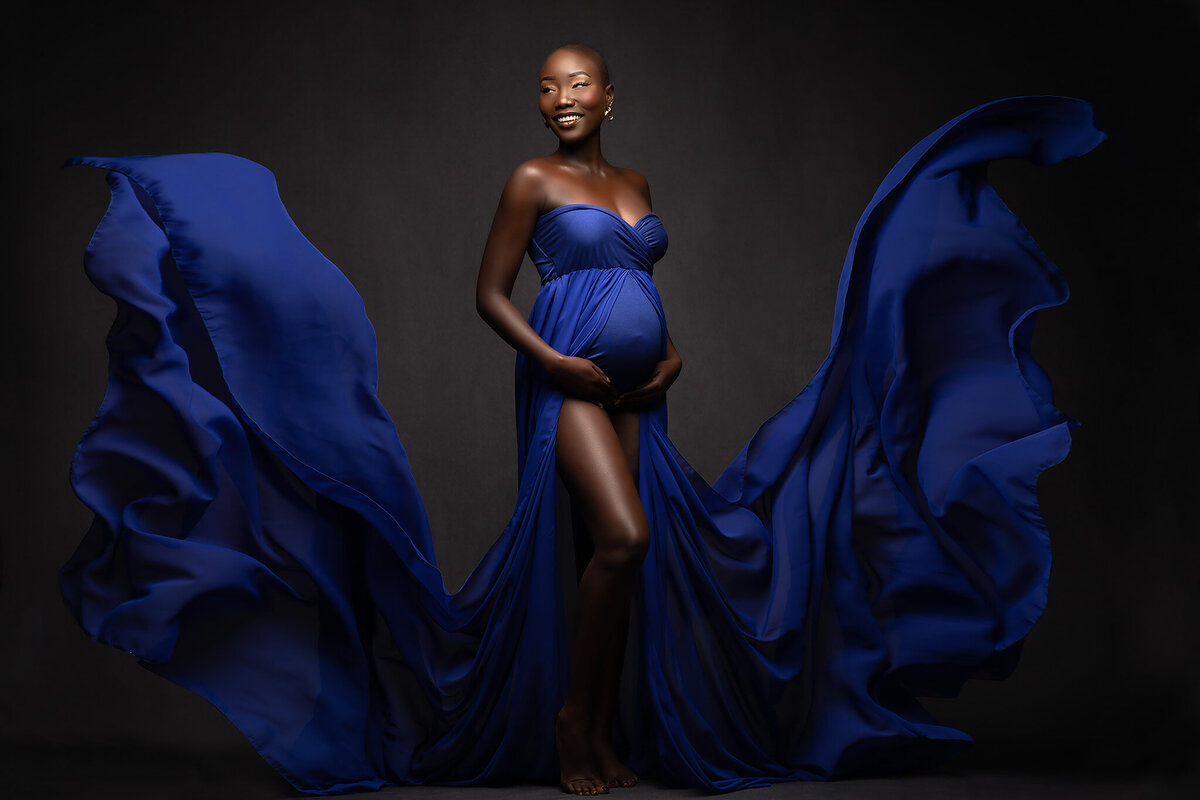 A radiant expecting mother stands confidently in a billowing royal blue gown that gracefully swirls around her. Her joyful smile and elegant pose capture the beauty of maternity photography in Dallas, showcasing the artistry of a skilled DFW maternity photographer.