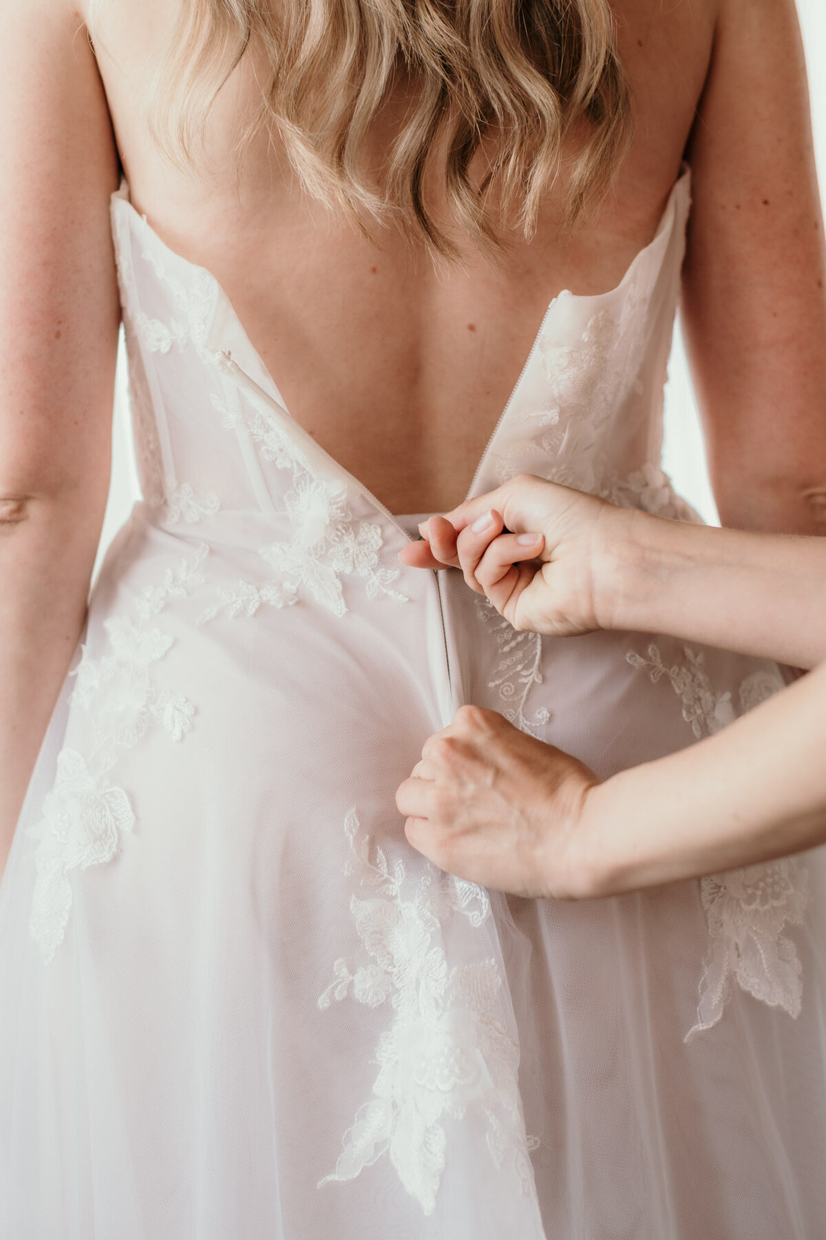Zwei Hände schließen den Reisverschluss des Brautkleids am Rücken der Braut.