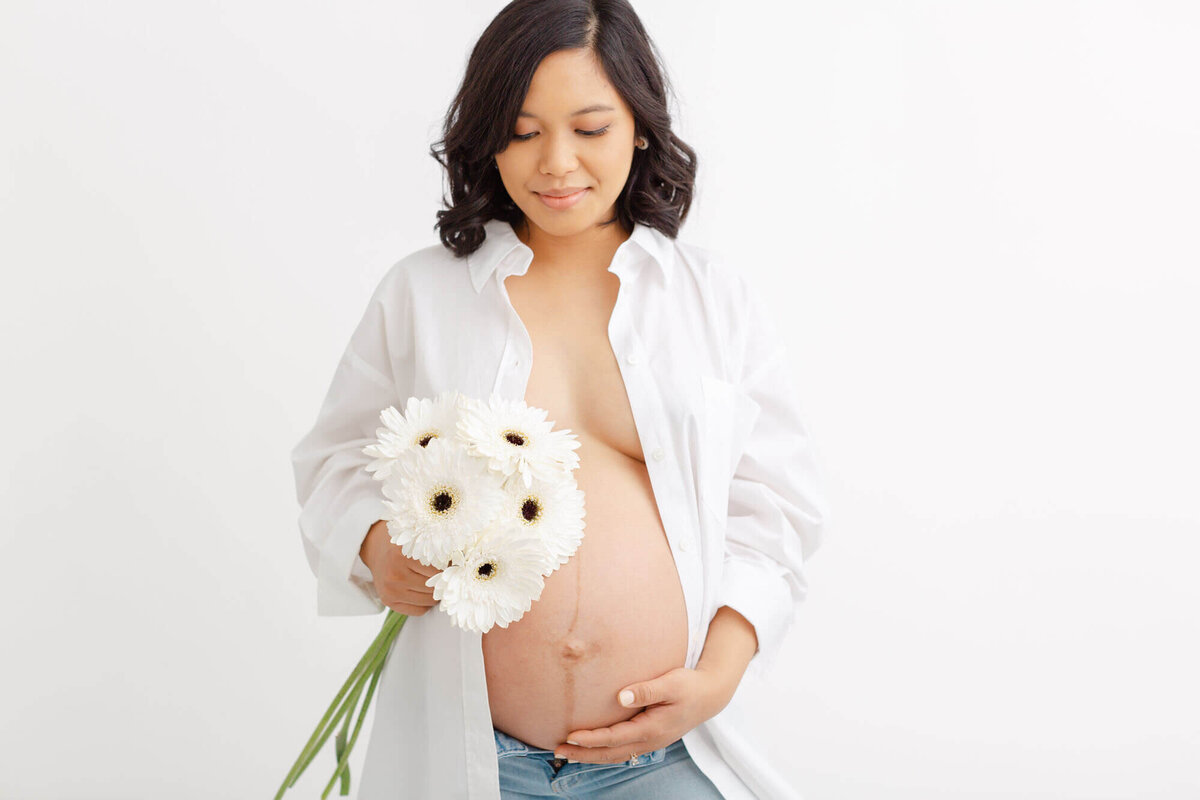 Woman with shoulder length dark wavy hair is wearing an opened button-down shirt showing off her bare pregnant belly. She is holding her belly on the bottom with one hand and holding white flowers at the top of her belly in the other hand.