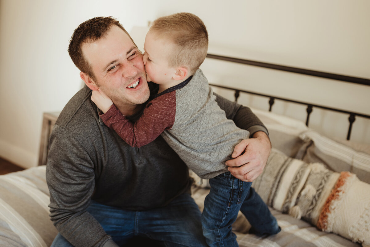 A little boy smooshes his face into his dad's cheek, giving him raspberries as his dad is laughing.