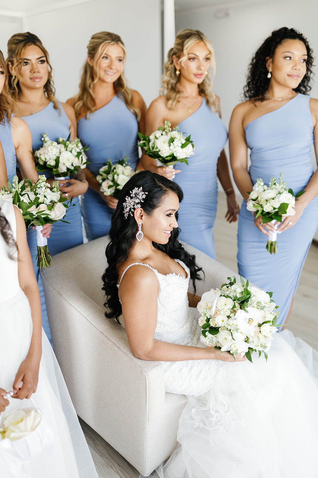 Bride Andressa sits elegantly with her bridesmaids in soft blue dresses, holding white floral bouquets, in a beautifully lit room at Table Key, Islamorada. Captured by Claudia Amalia, destination wedding photographer.