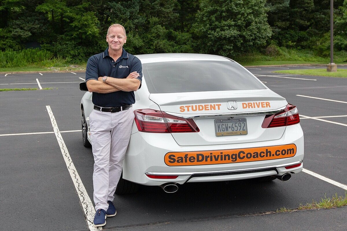 man leaning on a white car with the label Safe Driving Coach on the bumper.