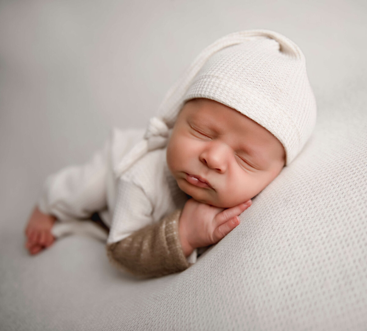 Side lying pose of a newborn baby boy in a white sleeper with sleepy hat in an Erie Pa photography studio