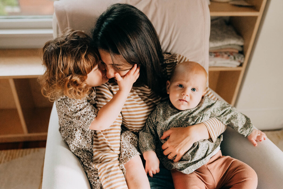 A toddler kisses her mother on the cheek while her mother holds her and her sister in a cream colored rocker in their nursery