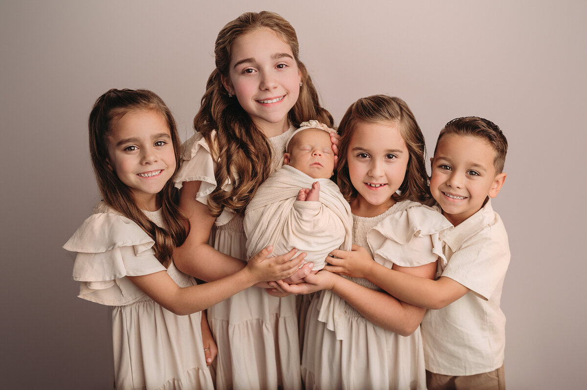 Stamford CT newborn photographer captures  newborn baby girl being held by her four older siblings whom are looking at the camera smiling