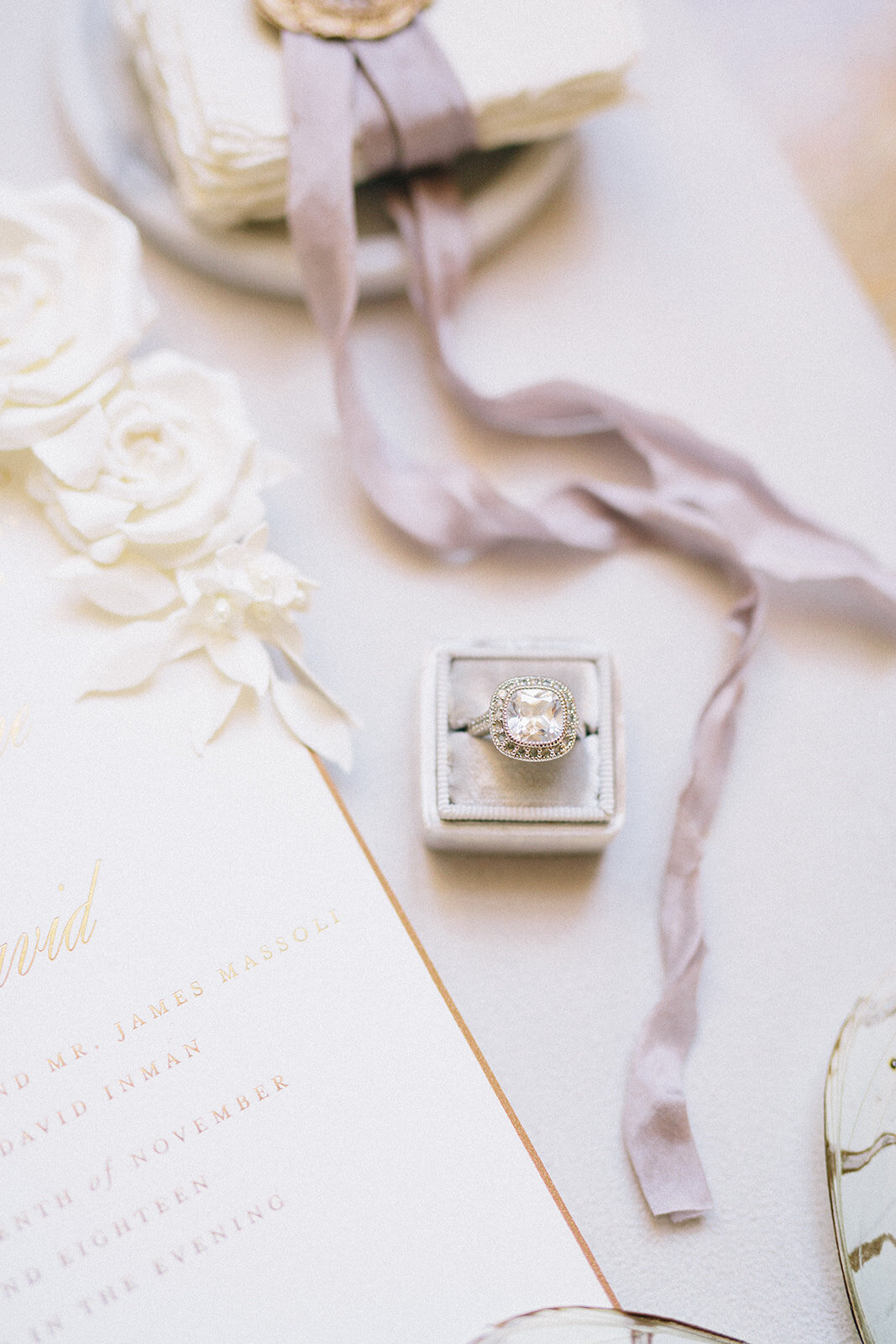 Close-up of a wedding invitation suite featuring a large diamond engagement ring in a velvet box, a wax-sealed envelope with purple ribbon, and white roses. The invitation text is partially visible, including names and a wedding date in gold lettering.