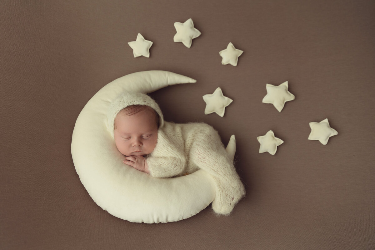 A newborn baby sleeps on a moon shaped pillow surrounded by matching stars in a knit onesie and beanie as posed for NJ Newborn Photography