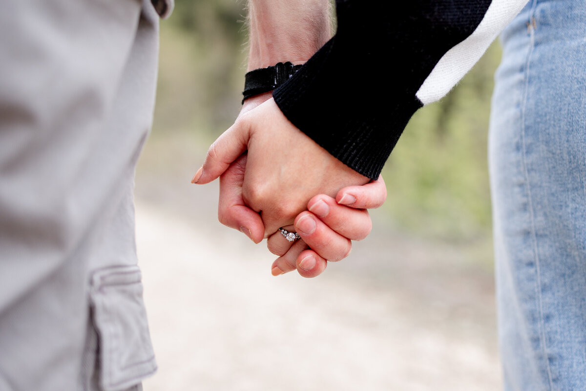 A close-up of a couple holding hands, featuring a beautiful engagement ring on the woman's finger against the blurred backdrop of a scenic trail.