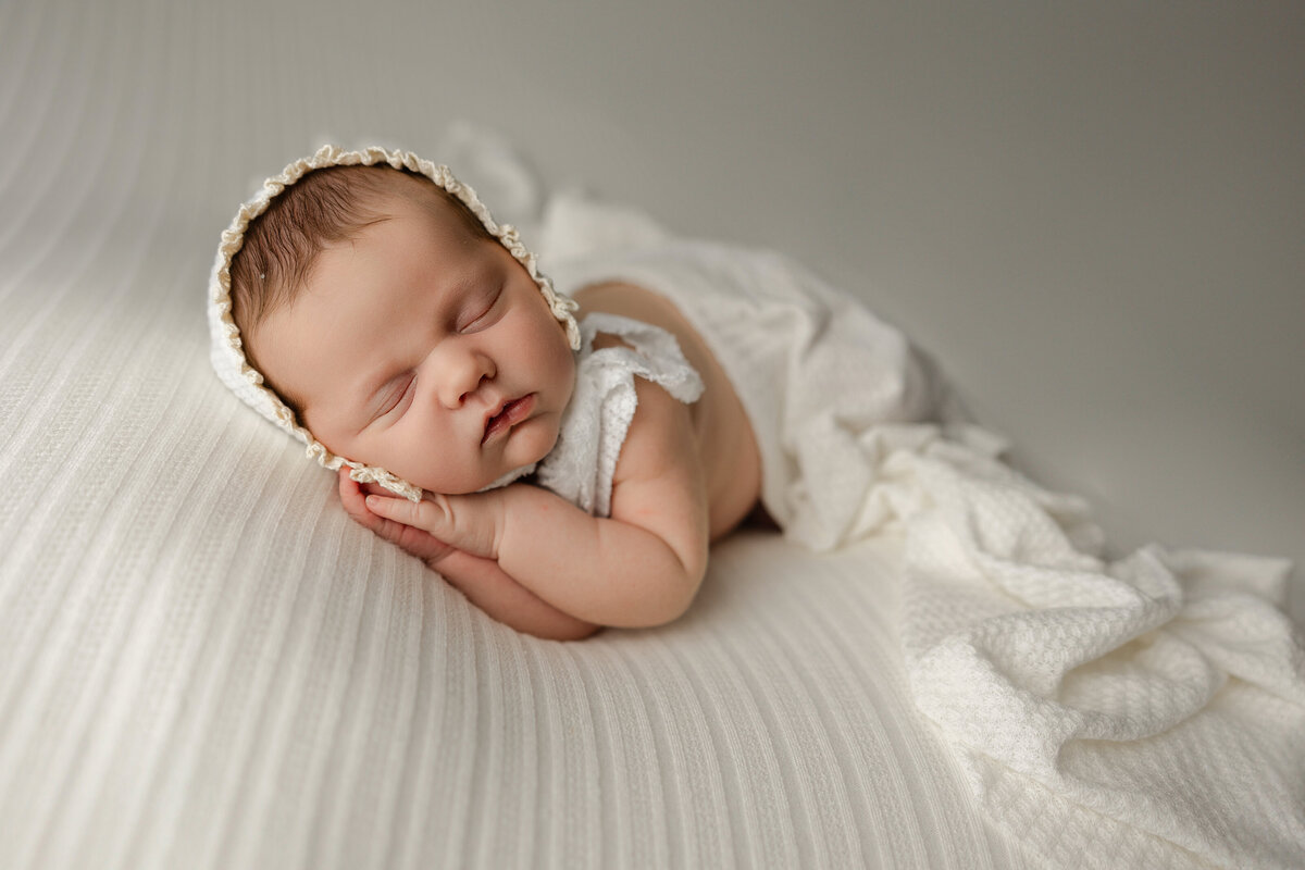 Newborn baby girl sleeping on a white textured blanket, styled with a lace bonnet and knit diaper cover during her studio session. Captured by Inland Empire newborn photographer Kaitlyn Dawn Photography.