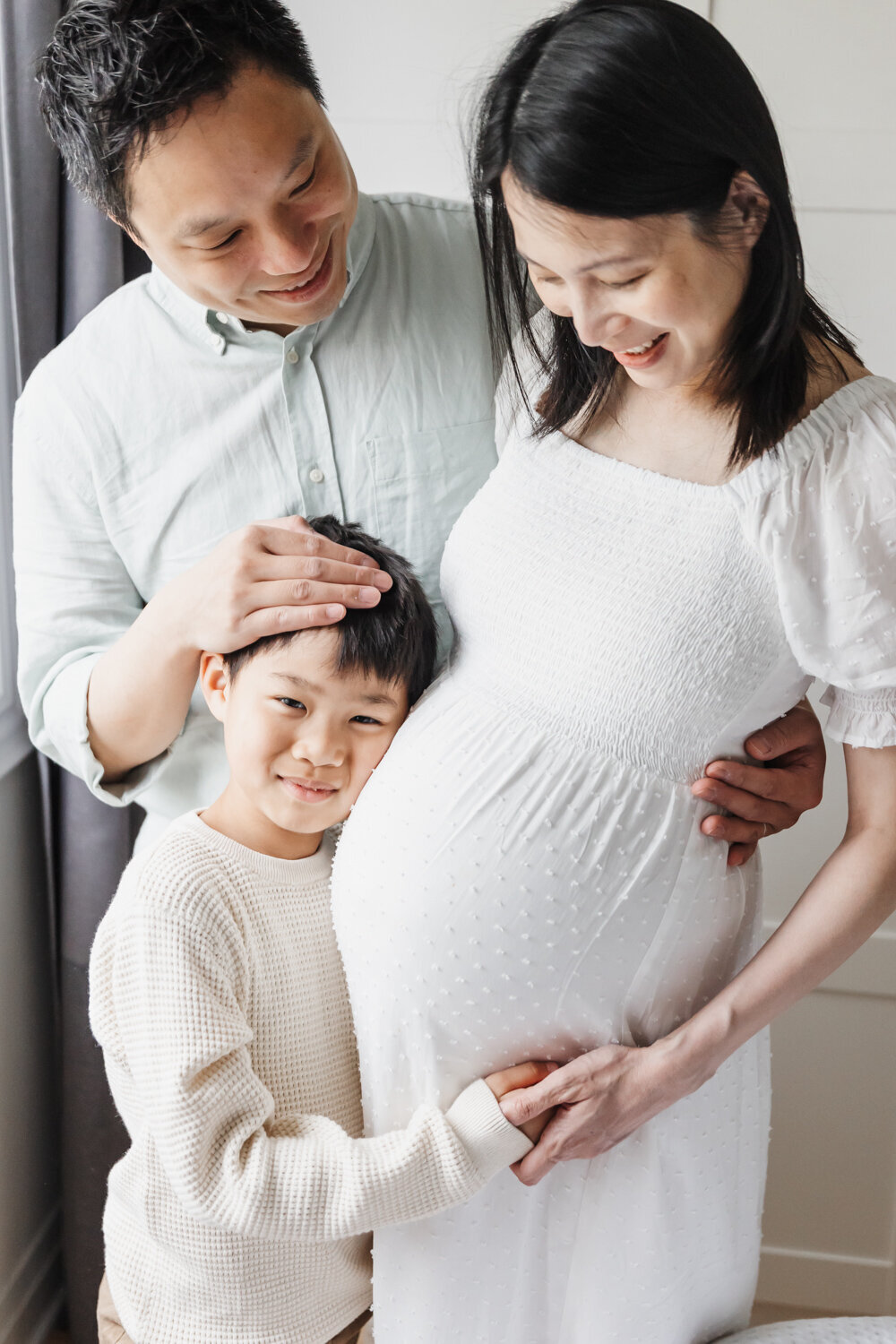 Older boy hugging his pregnant moms belly. Mom and dad are smiling down at him.