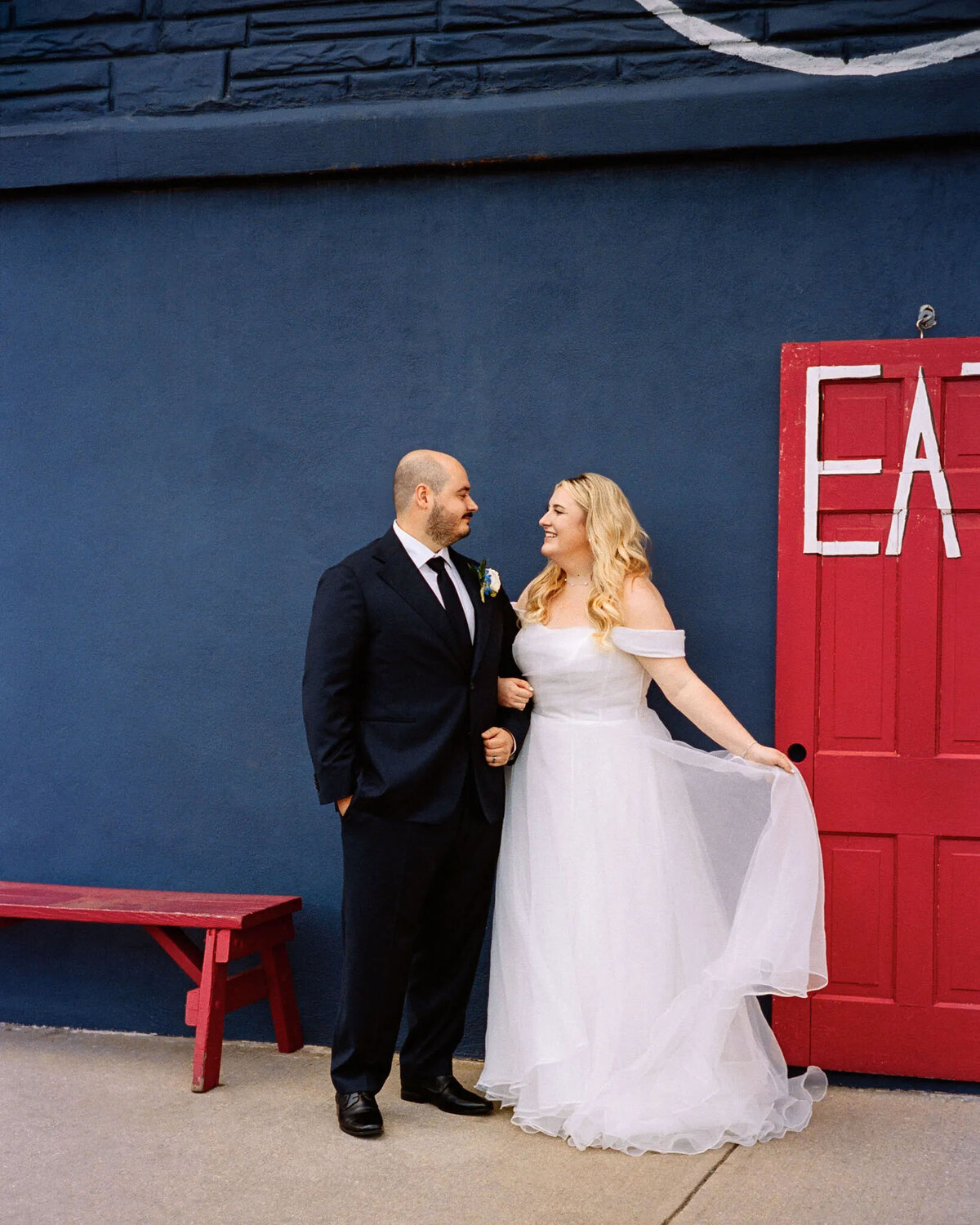 A bride in a white dress and groom in a dark suit smile at each other in front of a blue wall and a red door with EAT painted on it—a perfect moment for any film photographer NJ to capture. A bright red bench sits beside them.