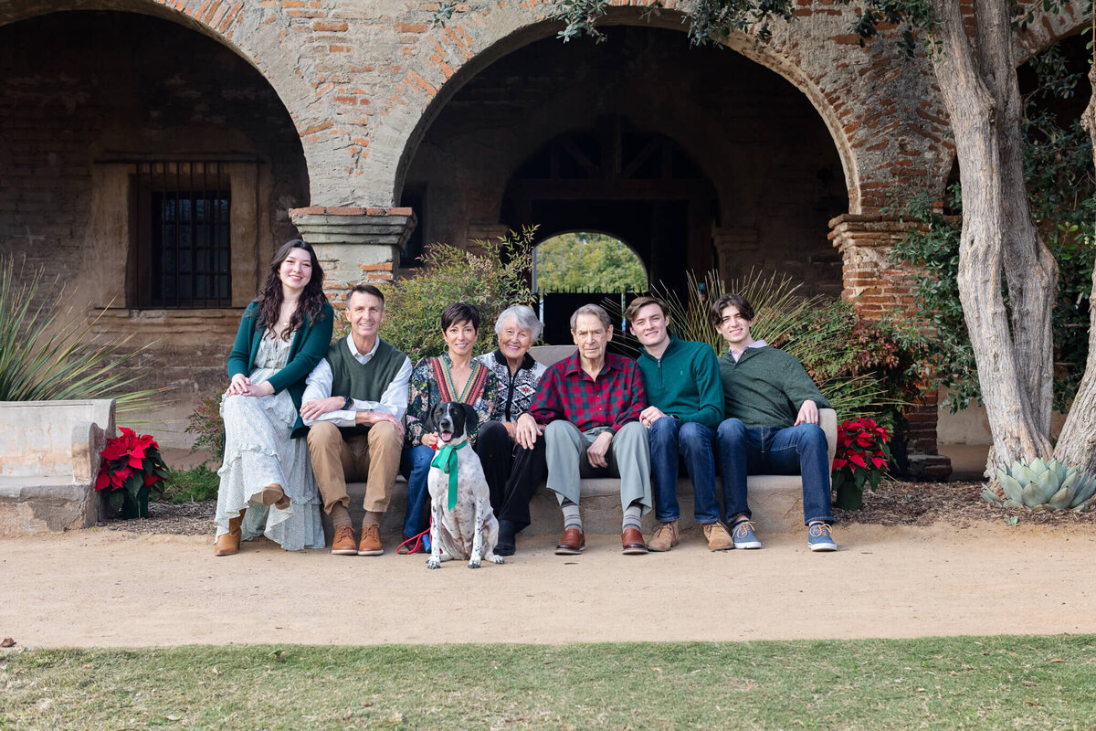 A large, extended family, sitting spread out on a bench beneath a brick archway looking at the camera.