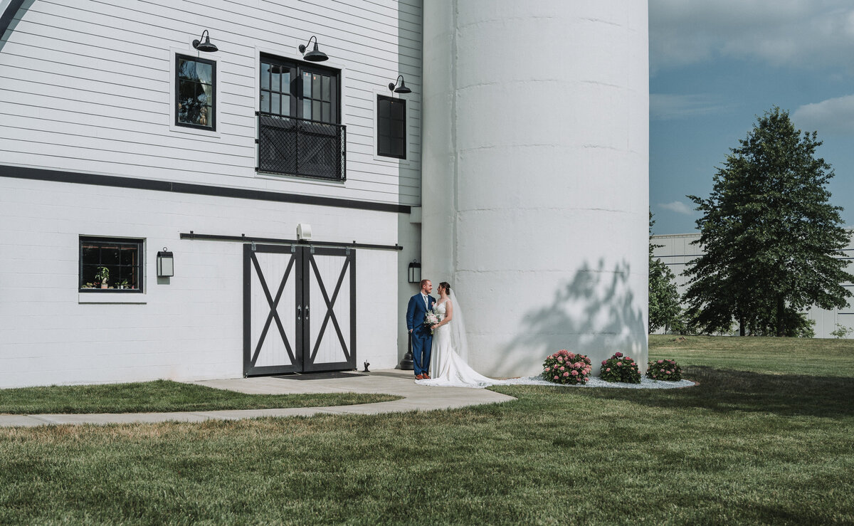 A newlywed couple embraces tenderly outside the iconic white barn and silo at Sweeney Barn in Manassas, Virginia, capturing a serene moment on their wedding day.