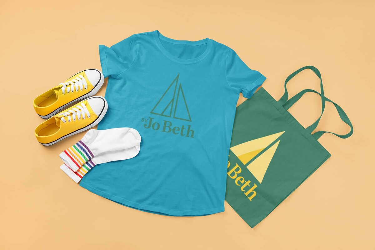 A blue t-shirt printed with a green SV Jo Beth one-color logo sits next to a green tote bag printed with a full-color yellow SV Jo Beth logo