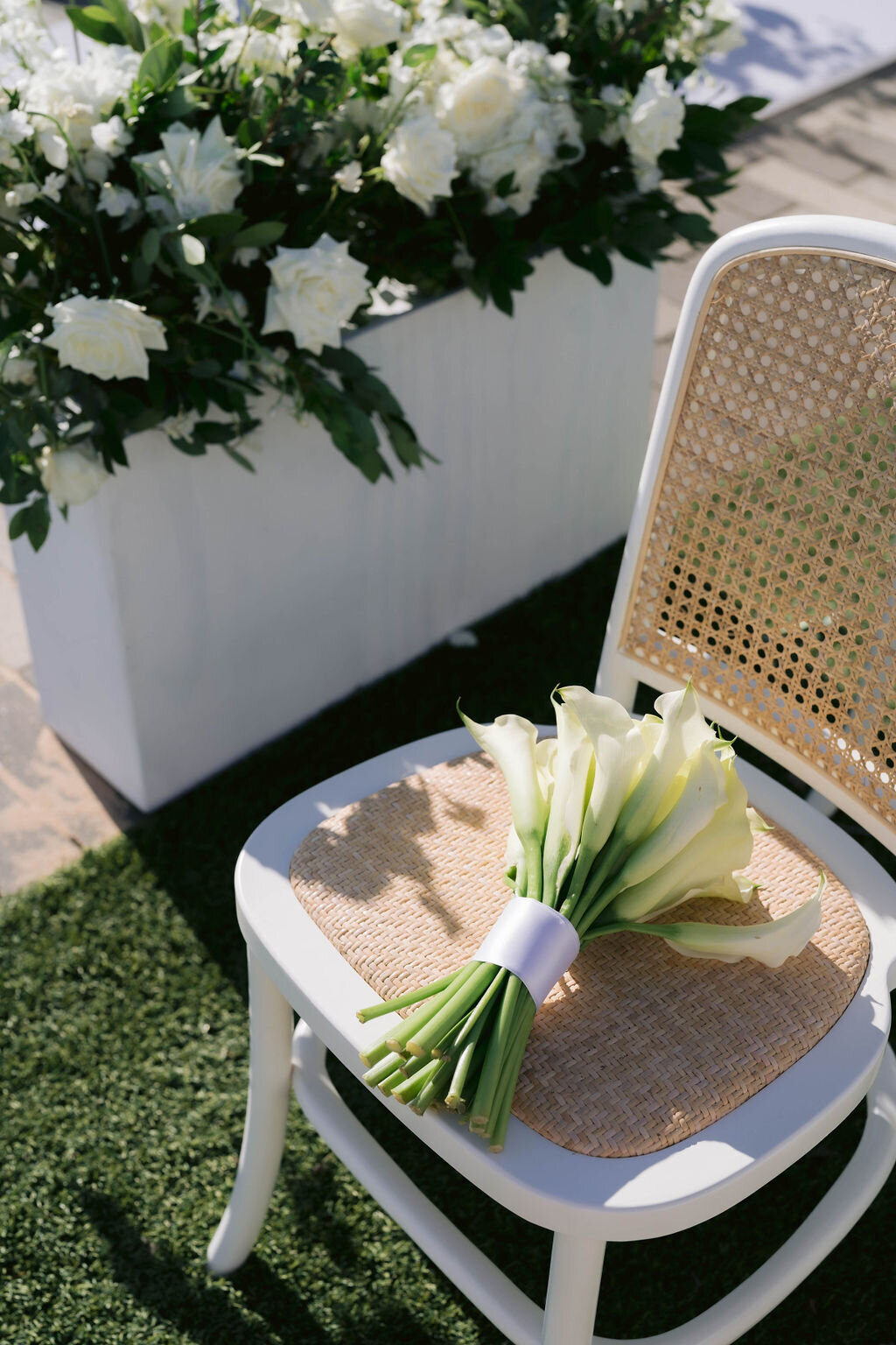 White cane-back chair with woven seat holding a calla lily bouquet beside a white floral planter at an outdoor Houston wedding.