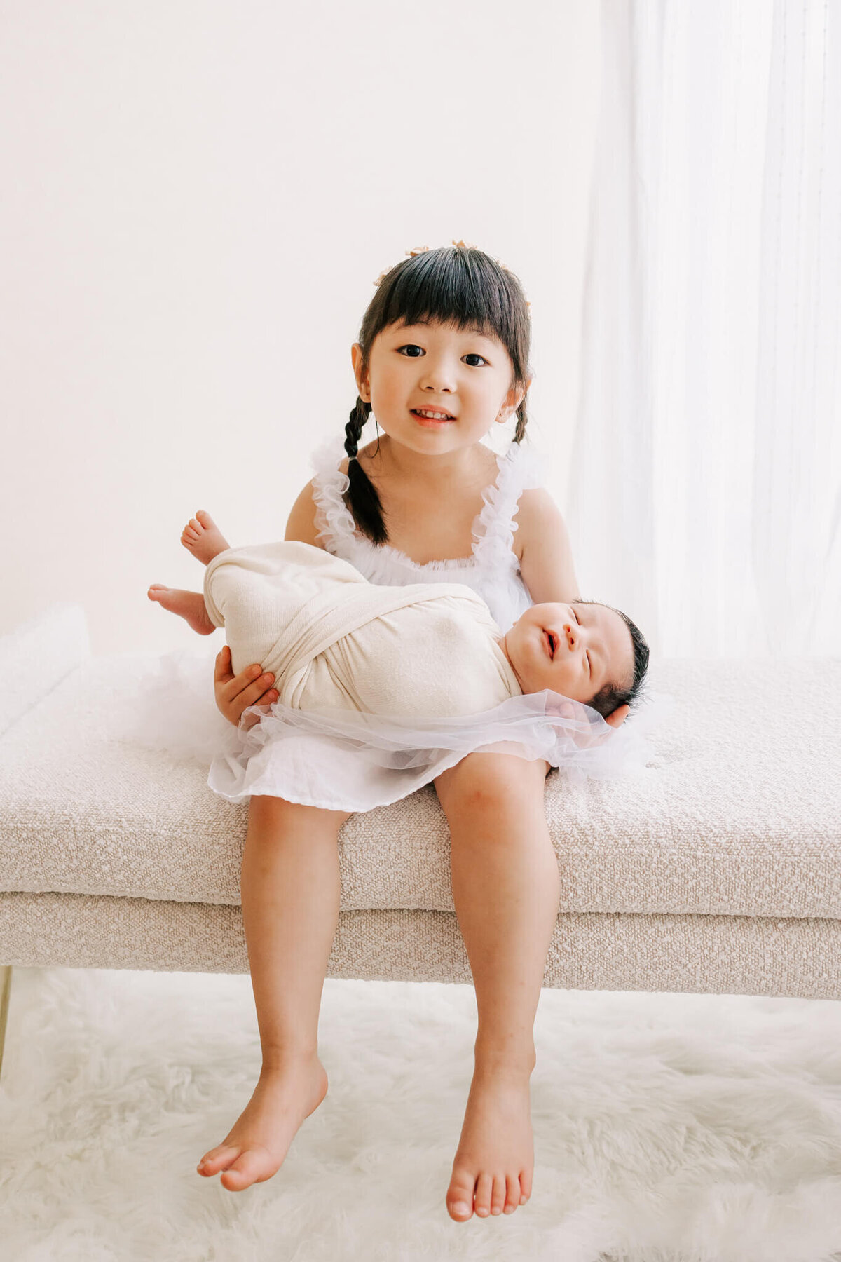 smiling older sister holding newborn baby brother on white bench