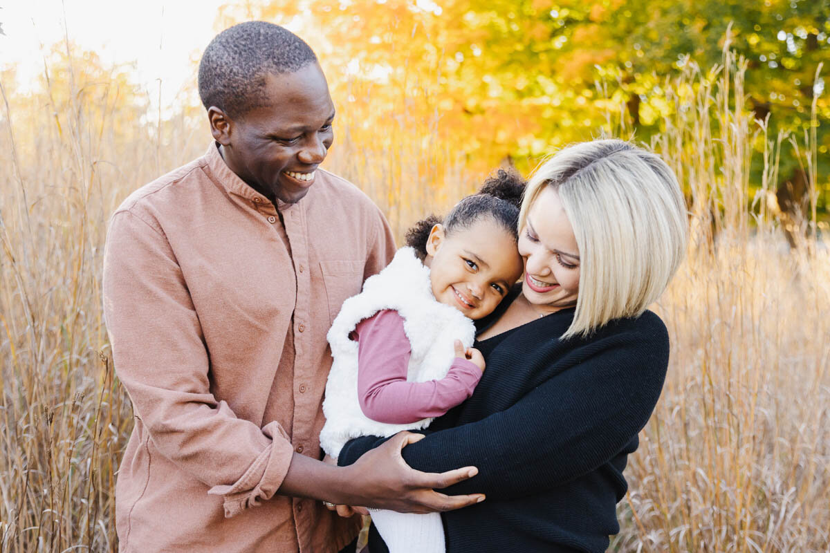 A man and woman stand in a field of tall grass, smiling and embracing a young girl between them, with autumn trees in the background—a perfect moment captured by a Toronto Family Photographer.