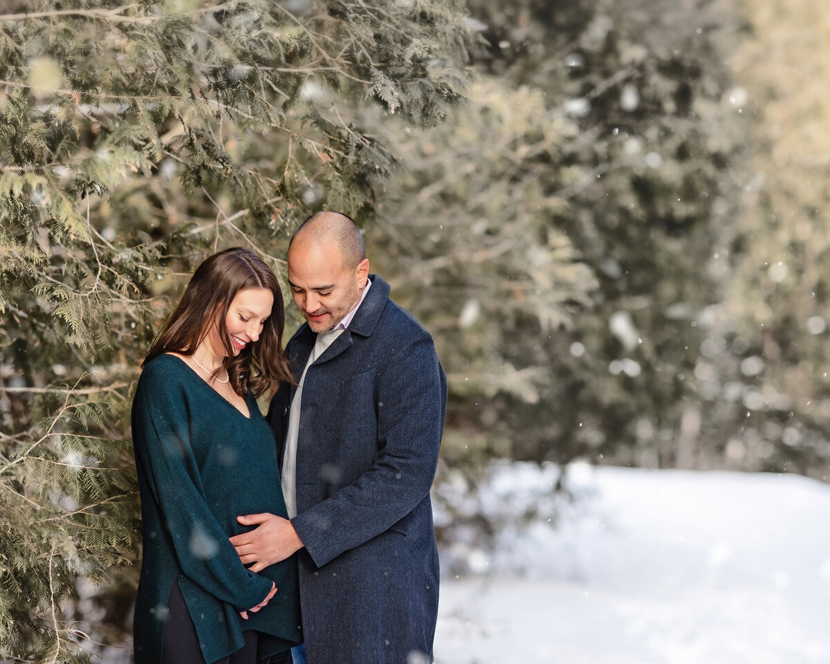 Expecting couple sharing a tender moment in a snowy pine forest during their winter maternity session in Paris, Ontario