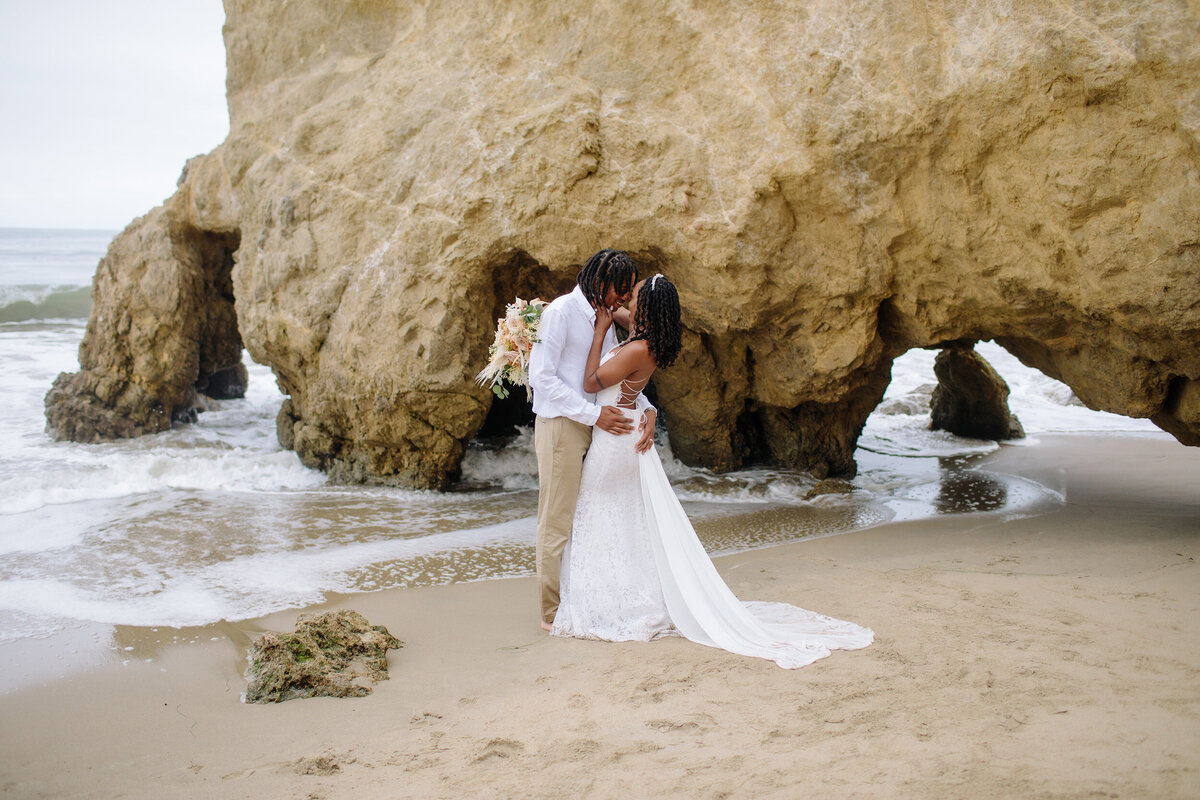 Bride and groom embrace with rock caves and the ocean in the background
