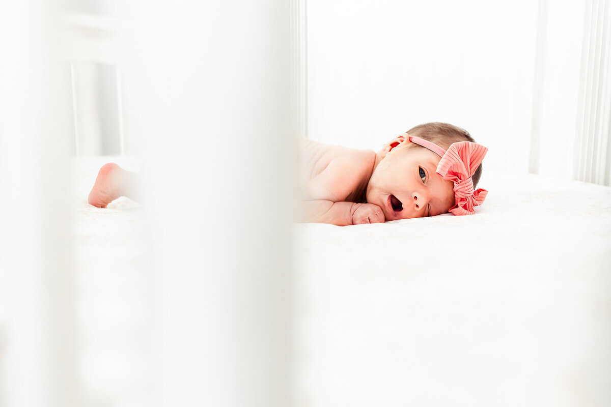 A newborn baby girl with a pink bow lays on her tummy in a bright white crib with light all around her.