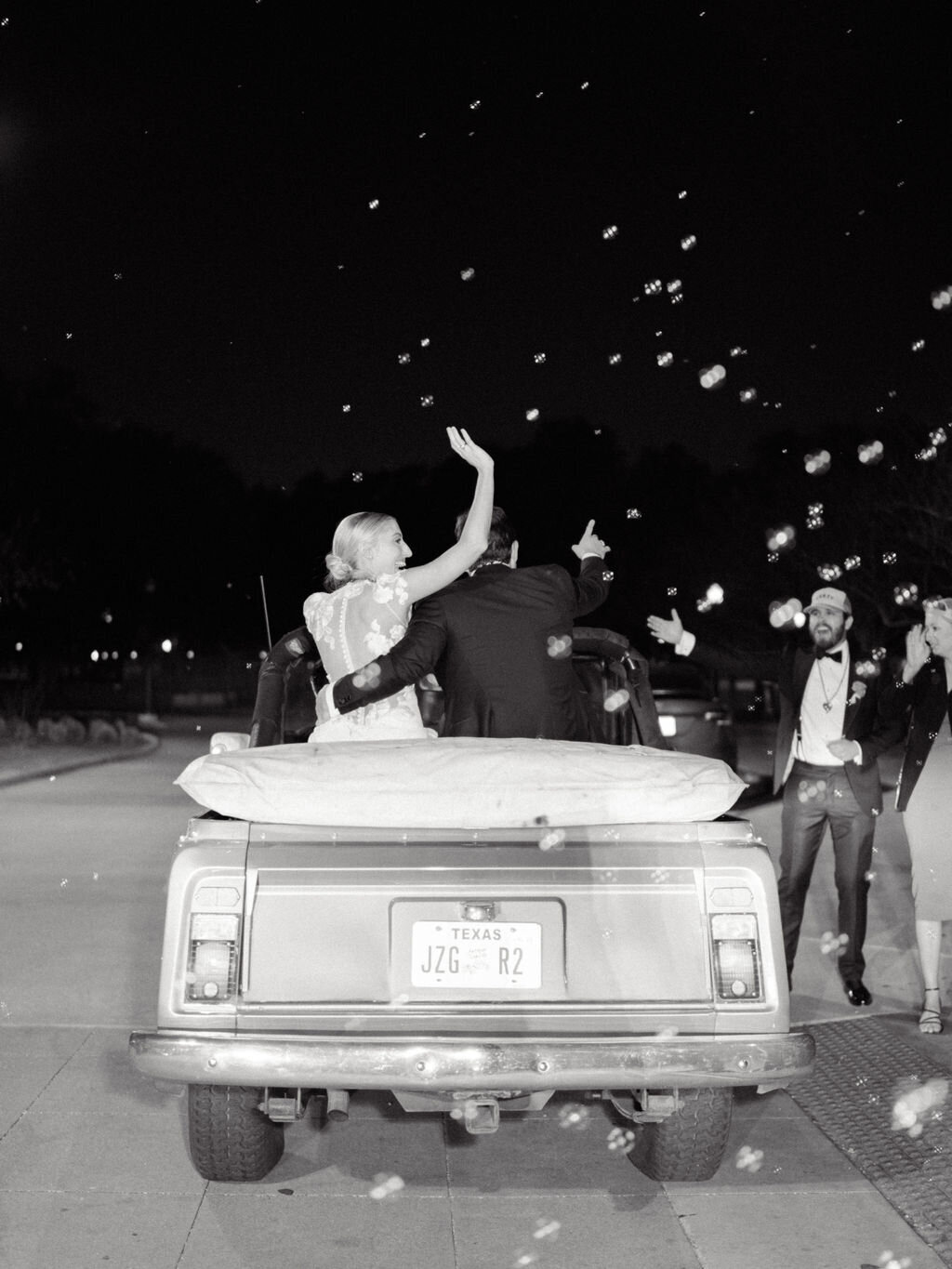 Black and white image of bride and groom waving in their getaway car surrounded by bubbles